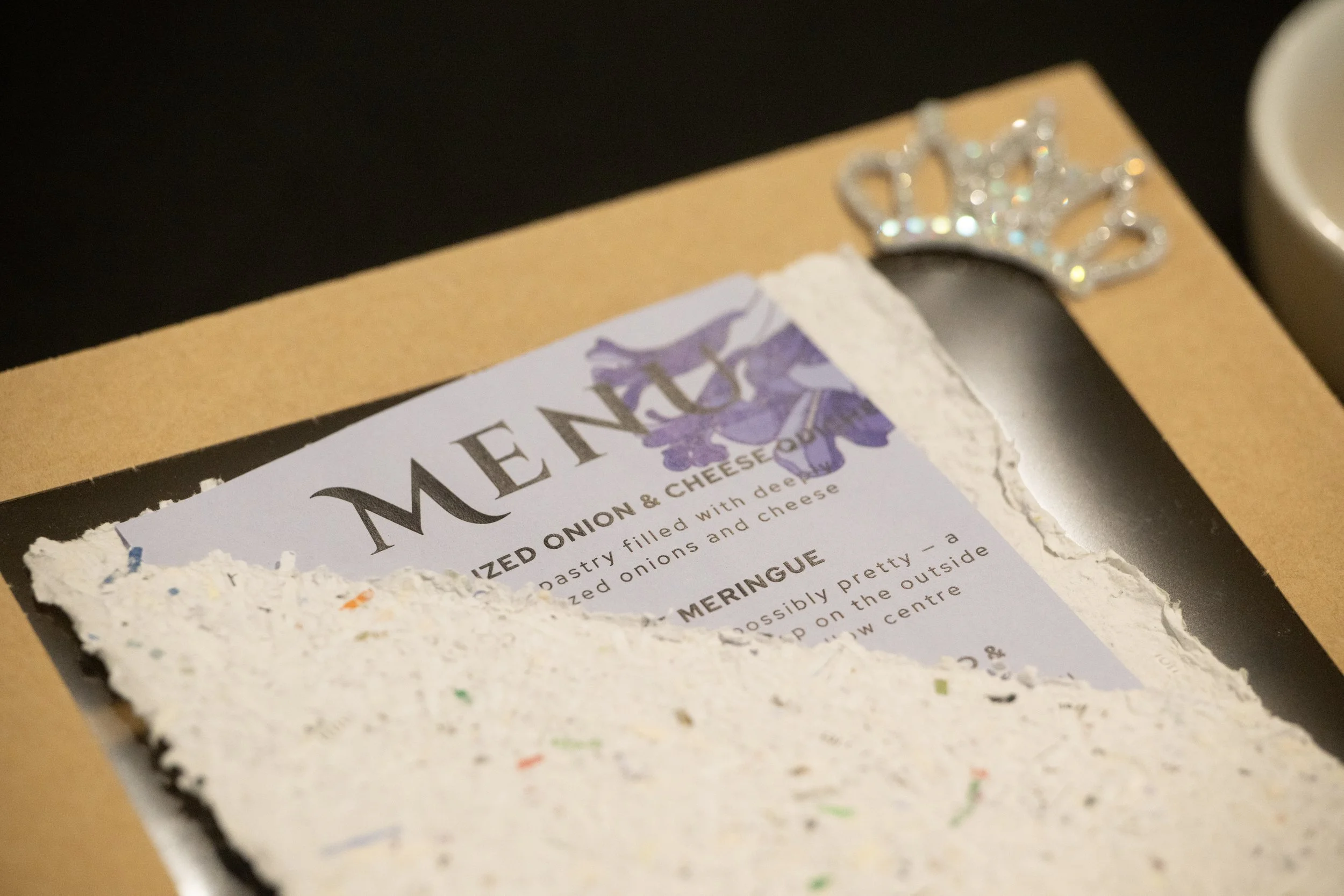 Close-up of a textured surface with torn paper and a portion of a recipe card titled "MEDITERRANEAN ONION & CHEESE" featuring a decorative rhinestone crown. The background shows a dark surface.
