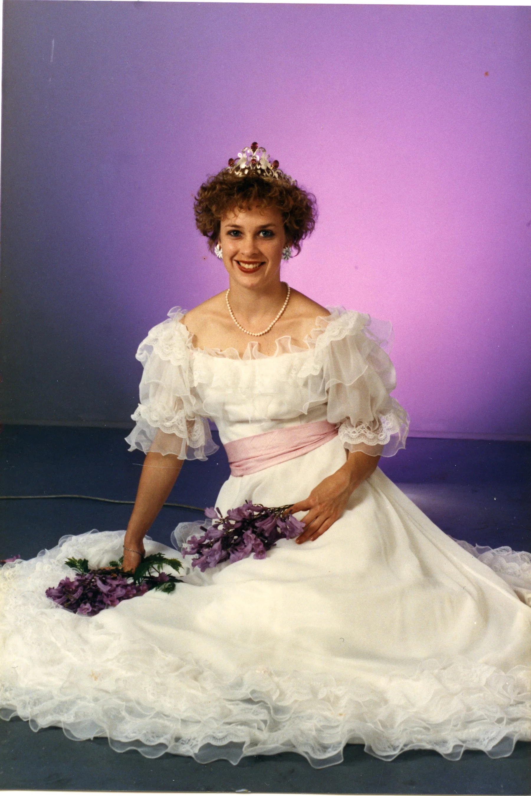 A woman dressed in a vintage white gown with ruffled sleeves and a pink sash, wearing a tiara, pearl necklace, and earrings, sitting in front of a purple background, holding purple flowers, with additional flowers on the floor.