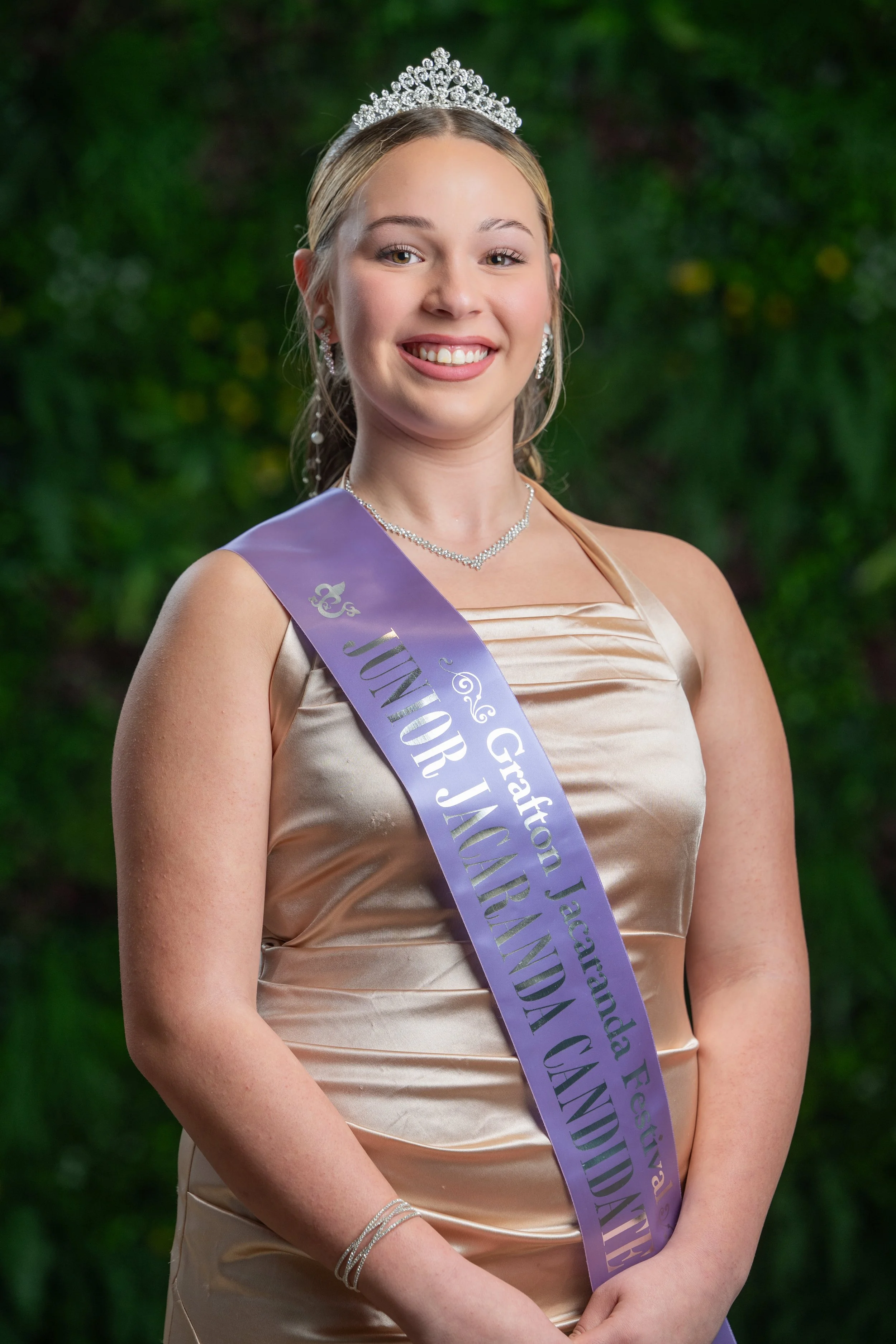 Young woman dressed in formal attire, wearing a sash that reads 'Junior' and a tiara, standing outdoors with a blurred green background.