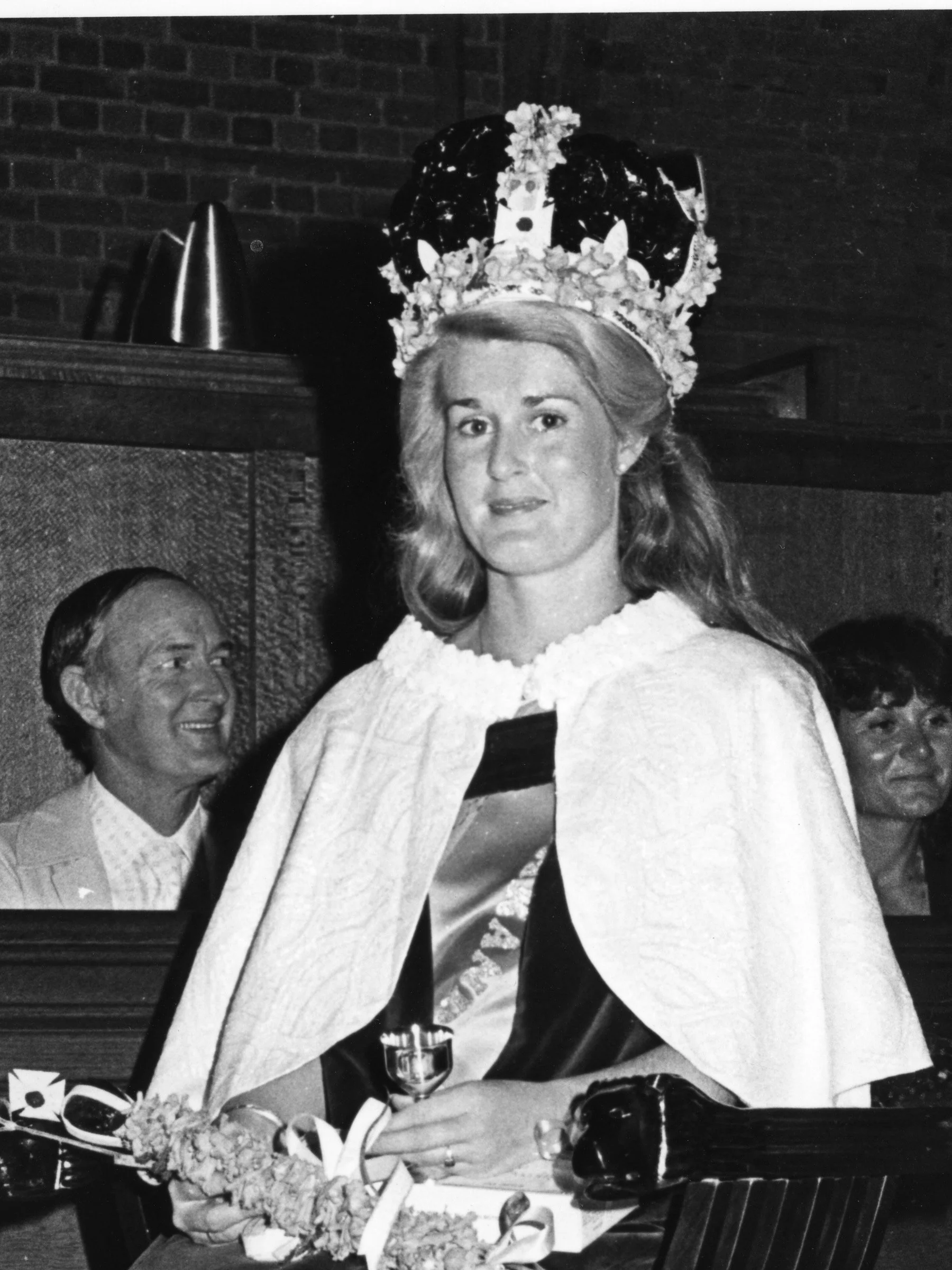 Black and white photo of a woman wearing a crown made of flowers, sitting at a table with food and drinks, smiling, with two men in the background.