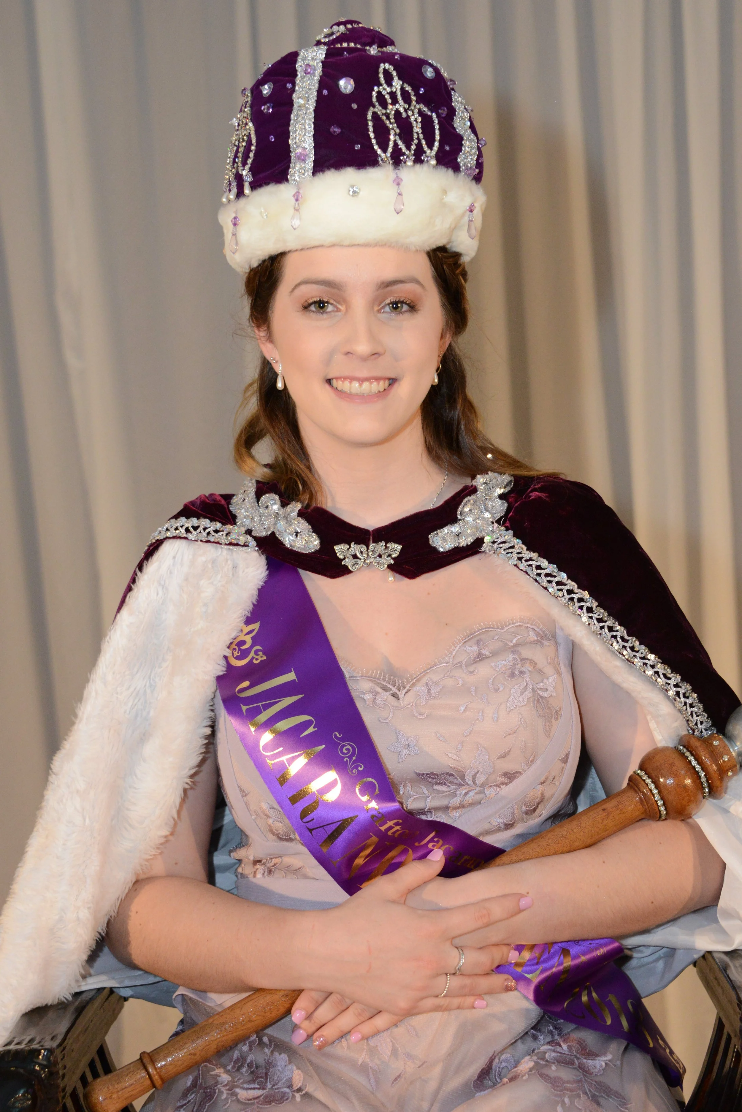 A young woman smiling, wearing a purple sash, a crown decorated with jewels and fur trim, a maroon cape with silver trim, and holding a scepter, indicative of a pageant or regal costume.