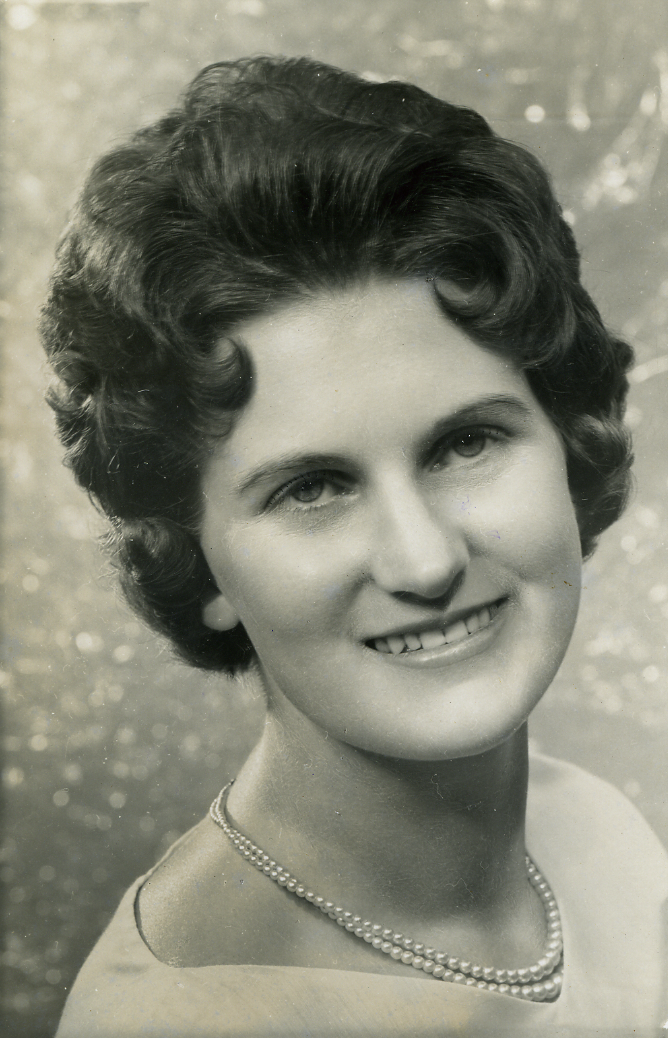Black and white portrait of a woman with short, wavy hair, smiling, wearing a pearl necklace and a light-colored top.