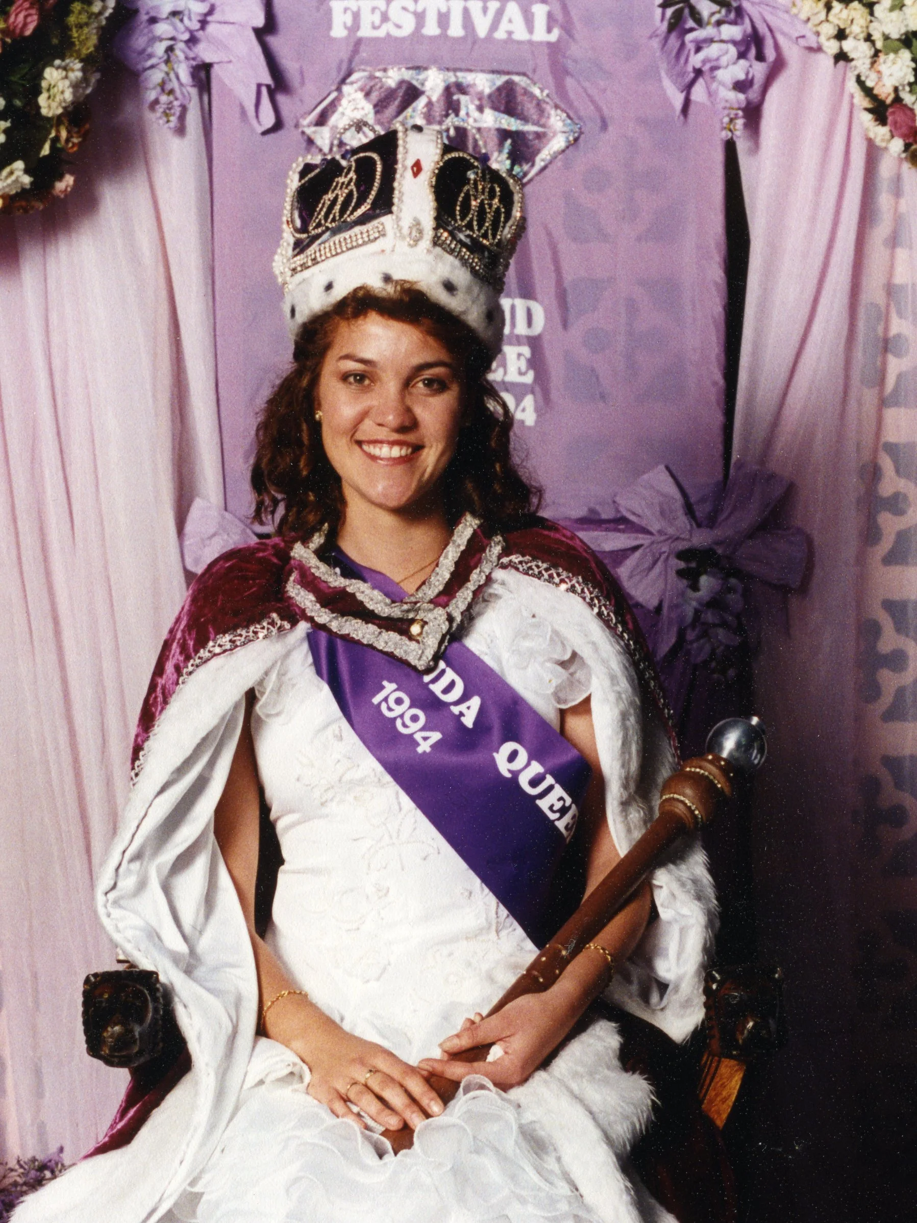 A woman dressed as a queen, wearing a crown and a sash, sitting on a throne with purple and pink decorations, holding a scepter.
