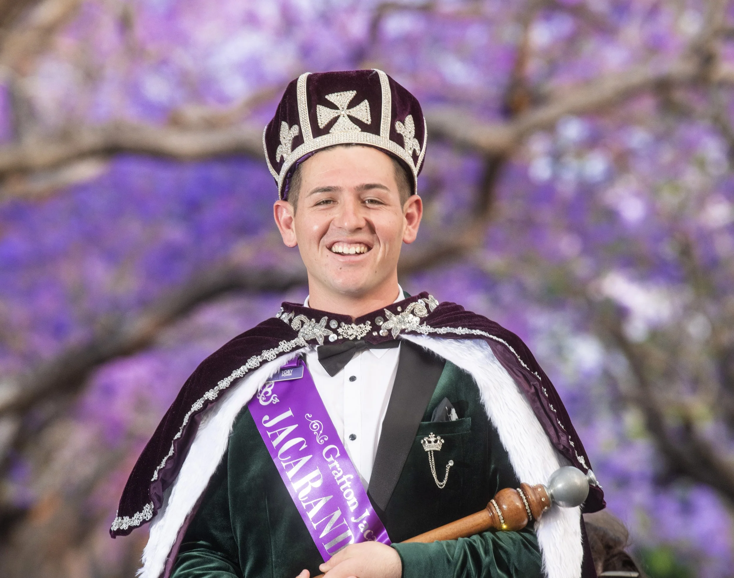 Young man wearing a crown, a velvet cape with silver embroidery, and a sash that reads 'Grand Duchess' smiling during a celebration surrounded by purple flowers in the background. He holds a ceremonial staff.