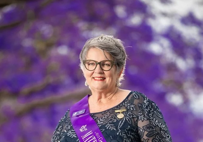 A smiling senior woman with short gray hair, glasses, and a tiara, wearing a purple sash with the title "Ms. [illegible]" and a black floral dress, standing outdoors in front of purple flowering trees.