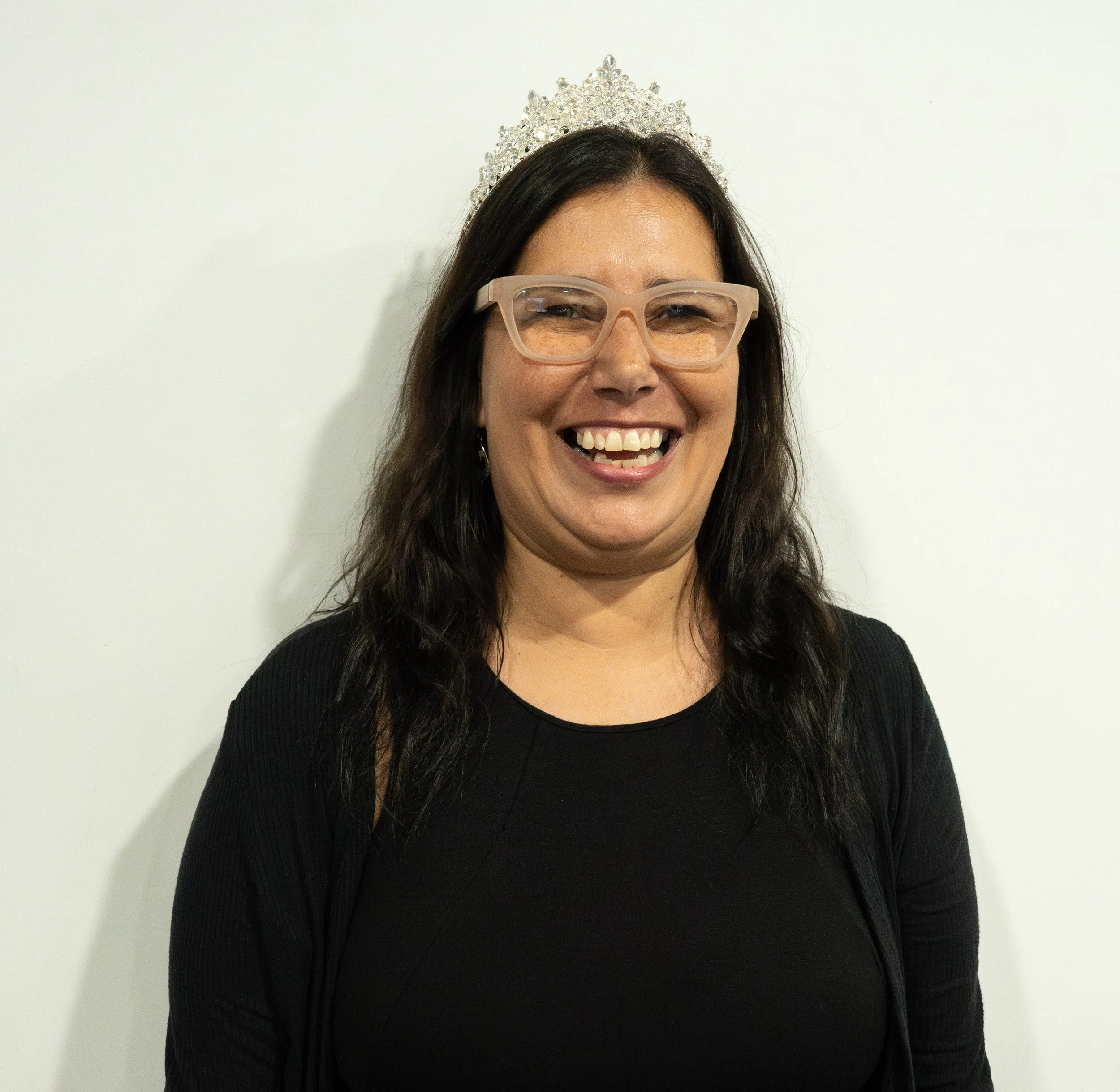A smiling woman wearing glasses and a tiara, with dark hair, standing against a plain white background.