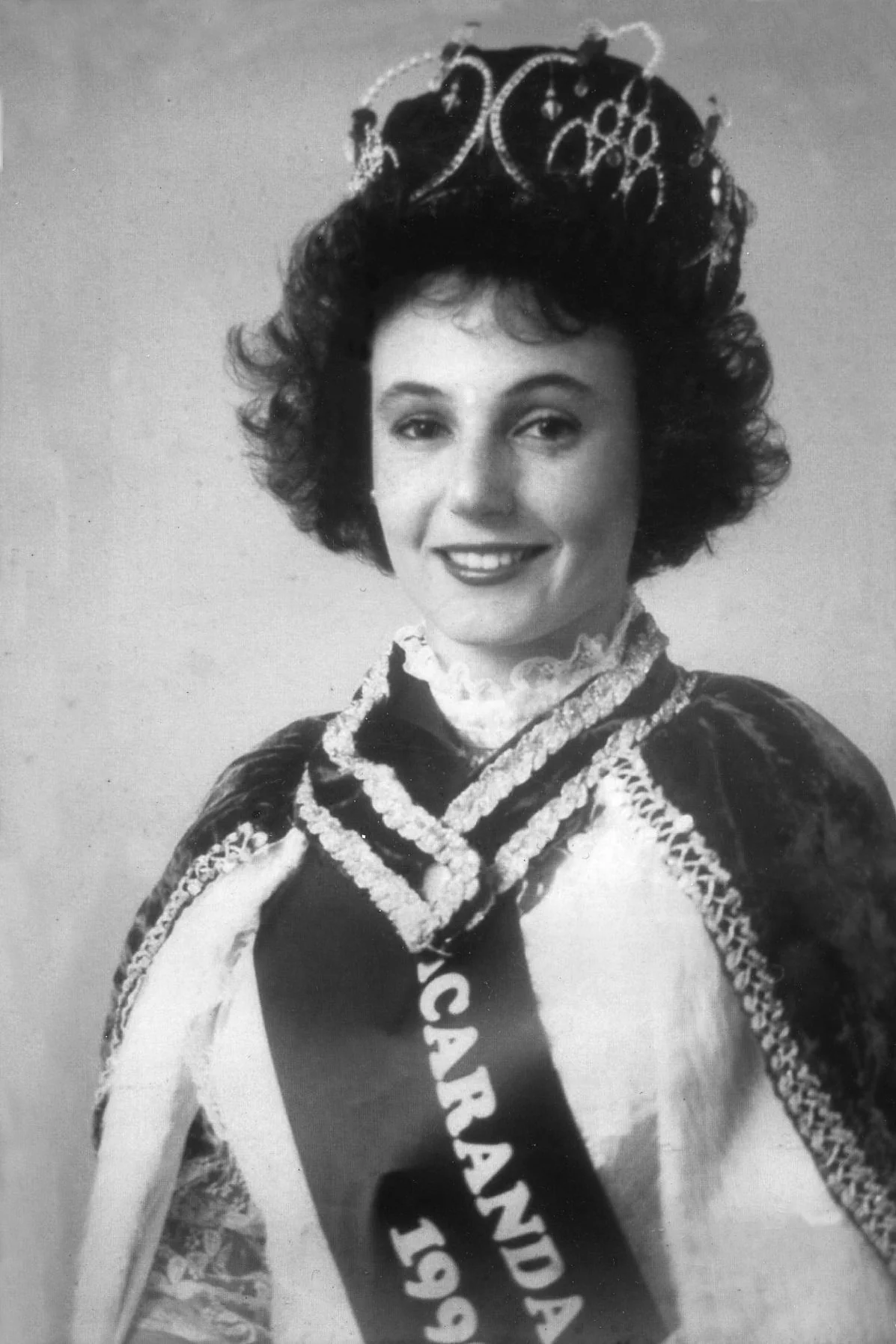 Black and white photo of a woman wearing a crown and a sash that reads 'California 1960'. She is dressed in an elaborate gown with lace and embroidery details, smiling at the camera.