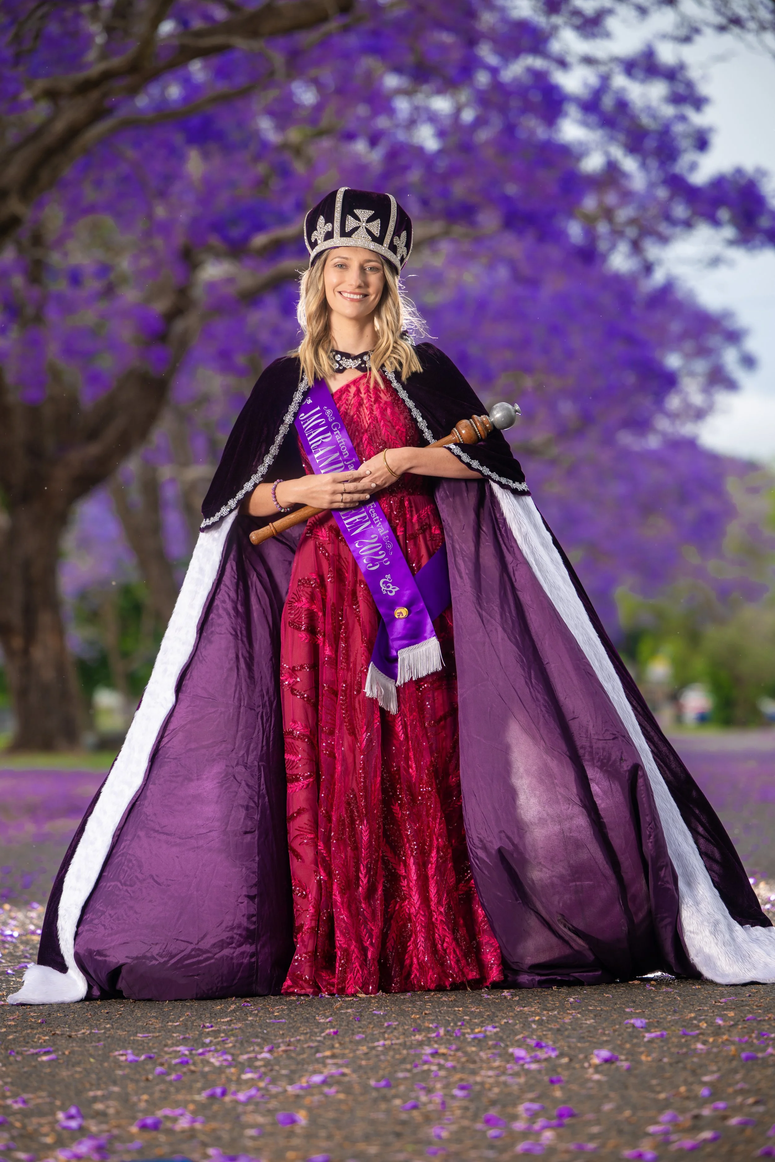 Woman in a purple and black queen costume with crown and scepter, standing outdoors near purple flowering trees.