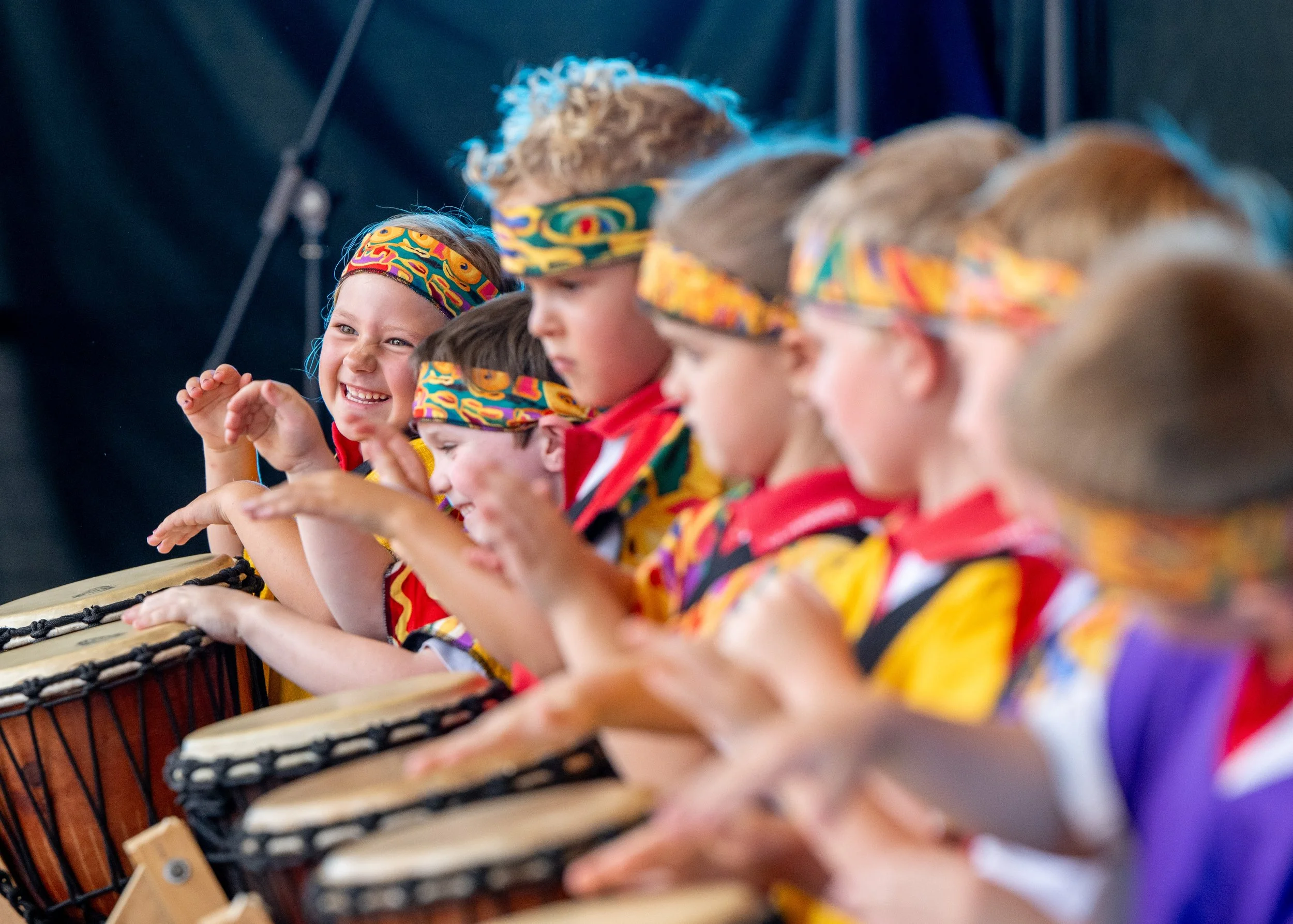 Children wearing colorful headbands and shirts, playing drums on stage during a performance.