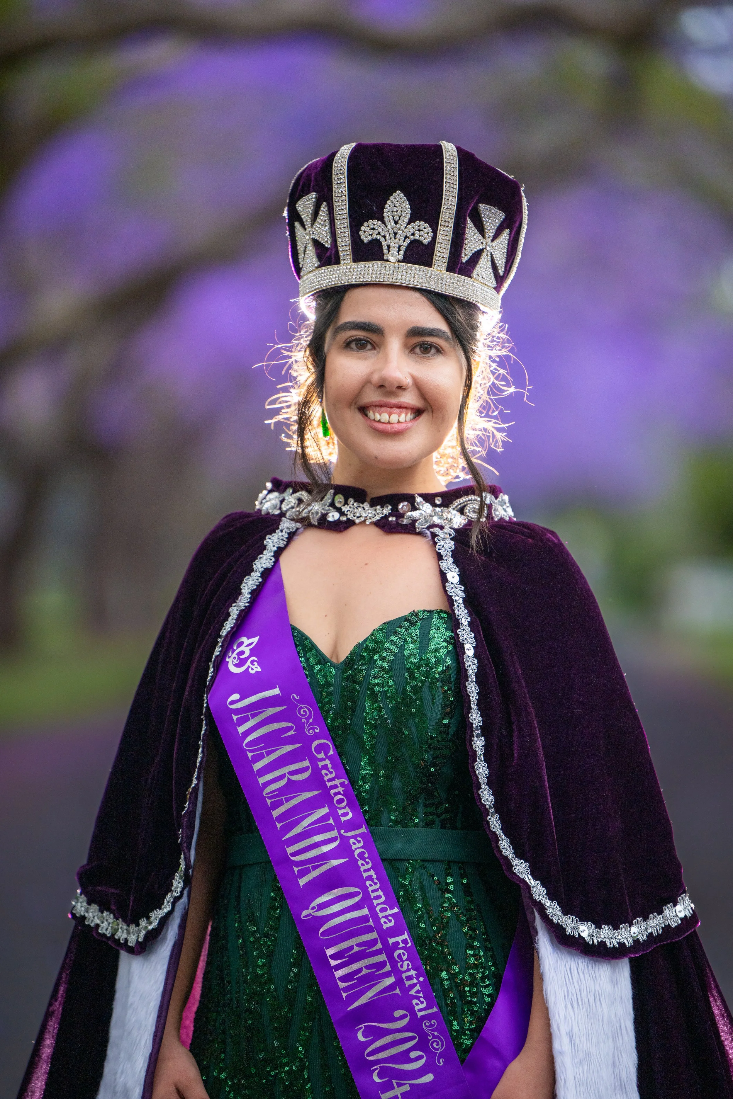 A young woman wearing a crown, a sash, and a velvet cape with silver trim, smiling outdoors with purple flowers in the background.