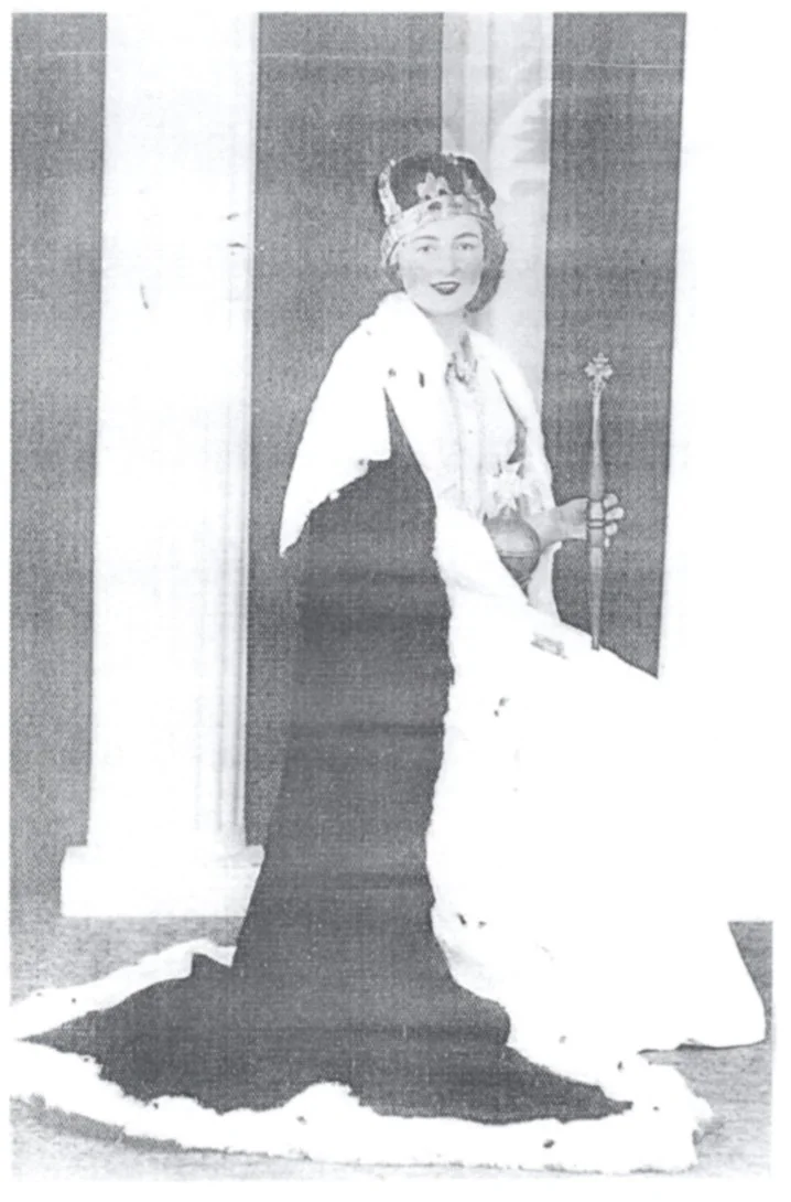 A vintage black-and-white photo of a woman dressed as a queen, wearing a crown, a long dark gown with a crown, holding a scepter, sitting on a chair with a white cloth draped over her shoulders.