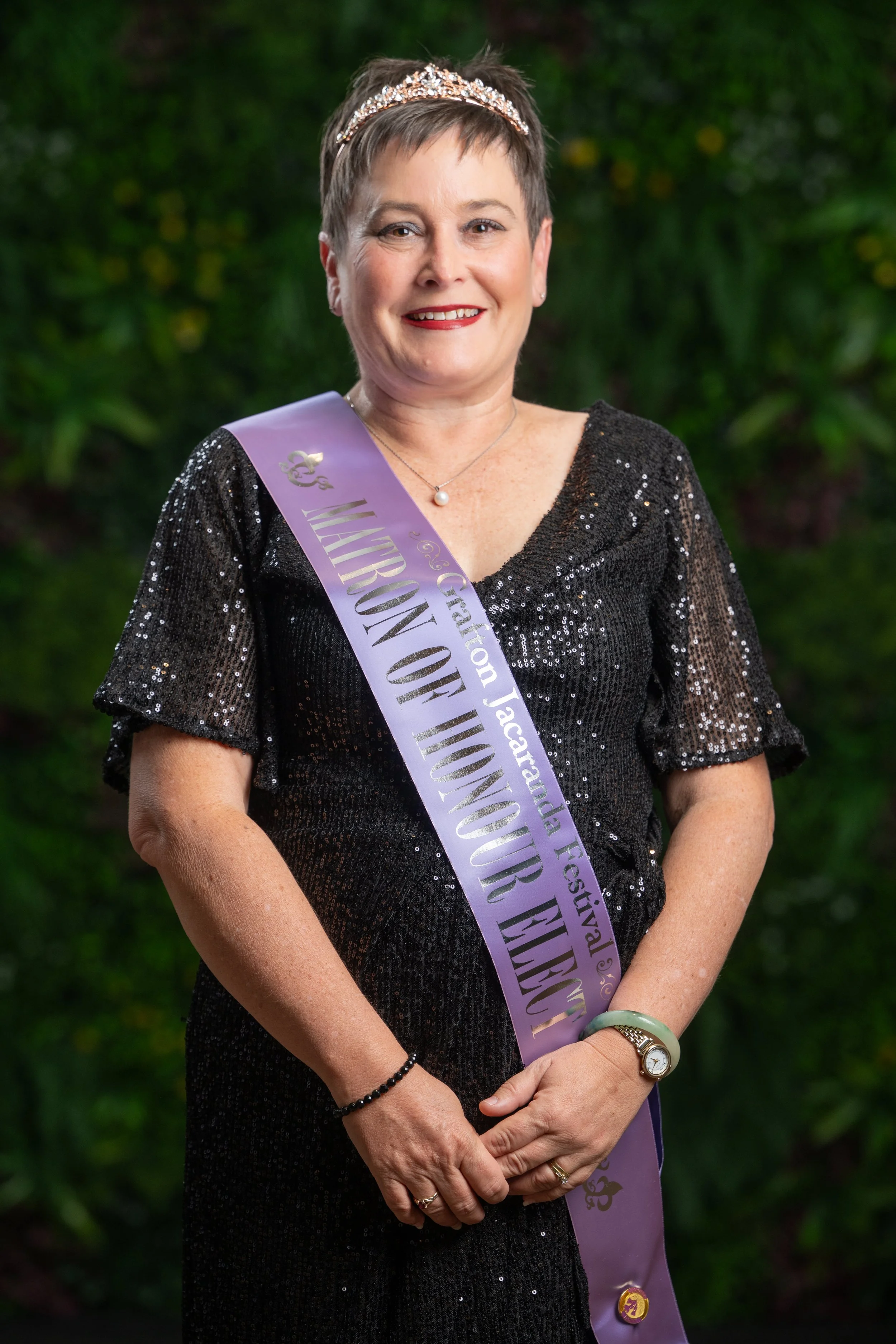 Smiling woman with short brown hair wearing a tiara, a black sequined dress, and a purple sash that reads 'Grefor 't National Jacaranda Festival' standing outdoors against a green background.