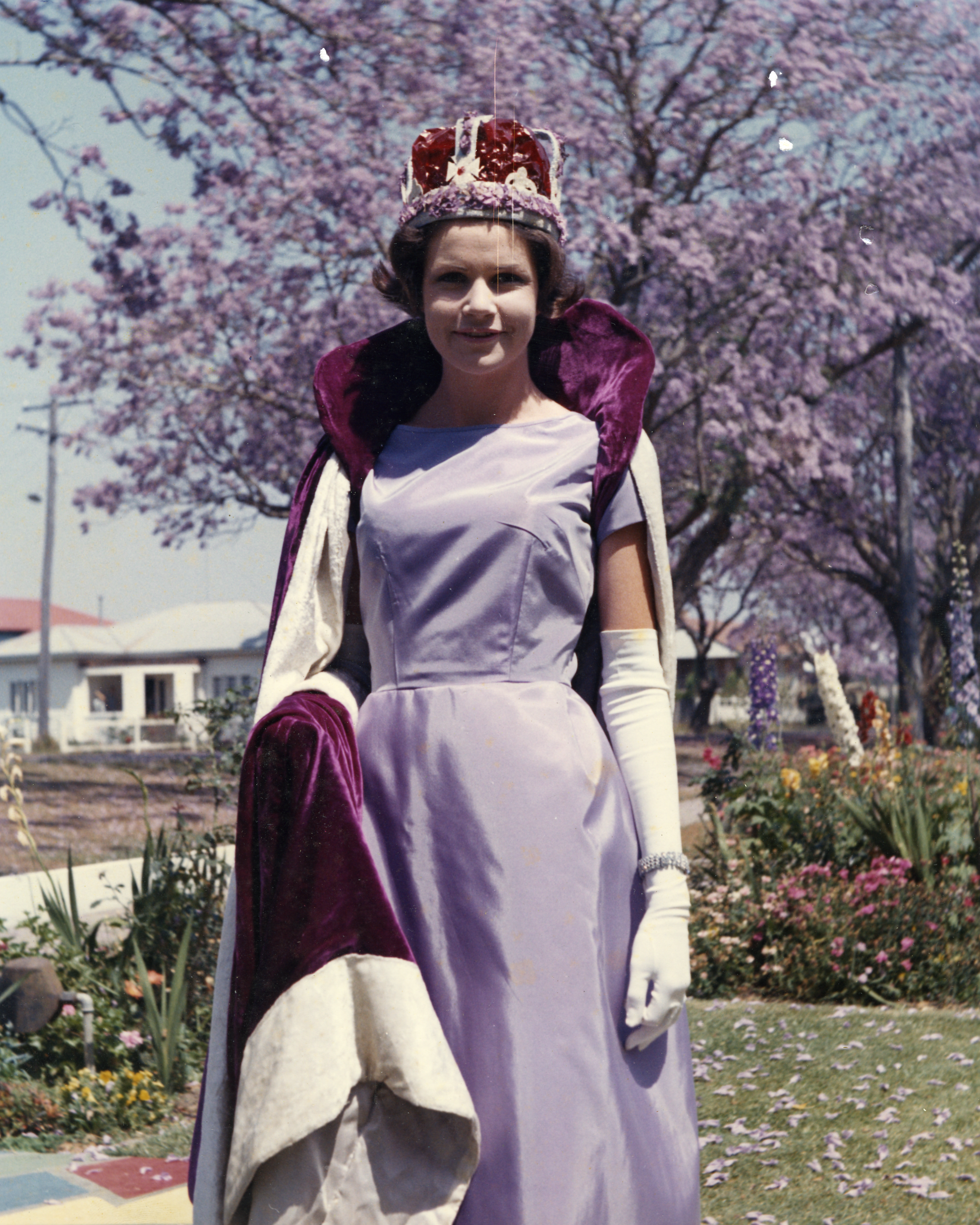 A young woman dressed as a queen or princess, wearing a crown and a purple cape, standing outdoors in front of blooming pink cherry blossom trees and a white building with windows.