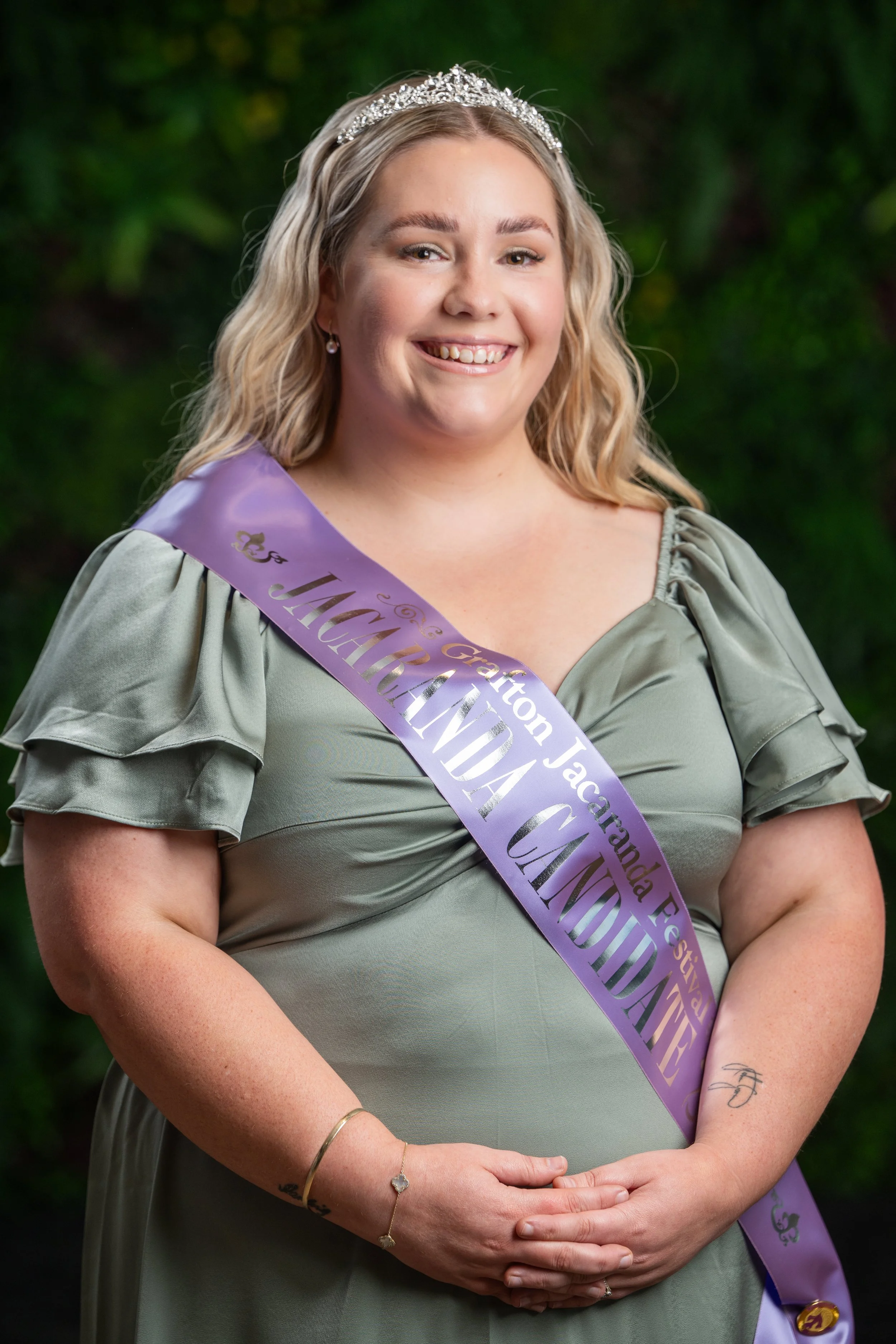 A woman wearing a tiara and a purple sash that reads 'JMU Hand' is standing outdoors against a background of green foliage. She is smiling, wearing earrings and bracelets, and has a tattoo on her left forearm.