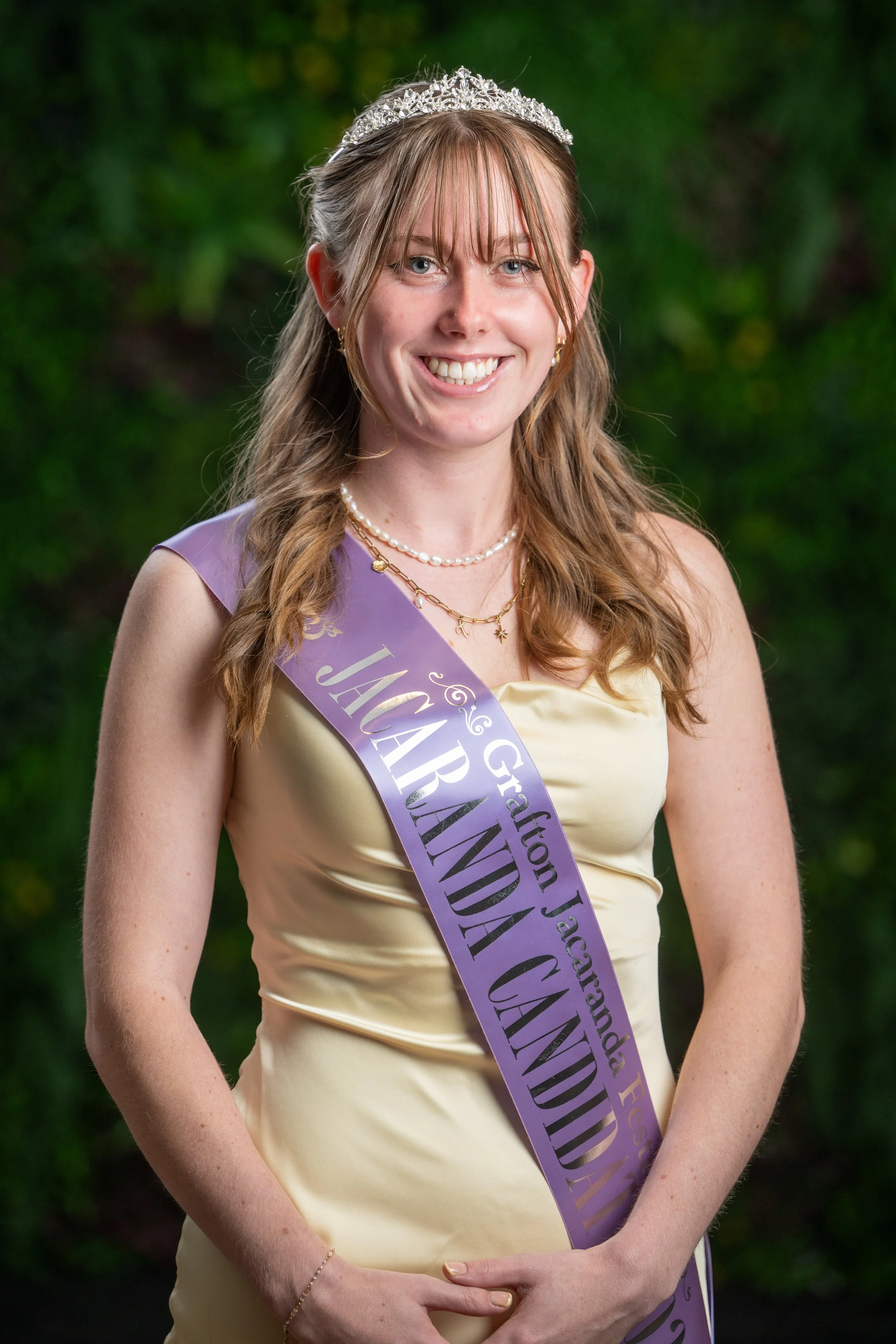 A young woman with long brown hair, wearing a tiara, a pearl necklace, and gold jewelry, smiling at the camera in an outdoor setting with green foliage background. She has a purple sash with gold lettering that reads 'Granddaughter' in gold and other words partially visible.
