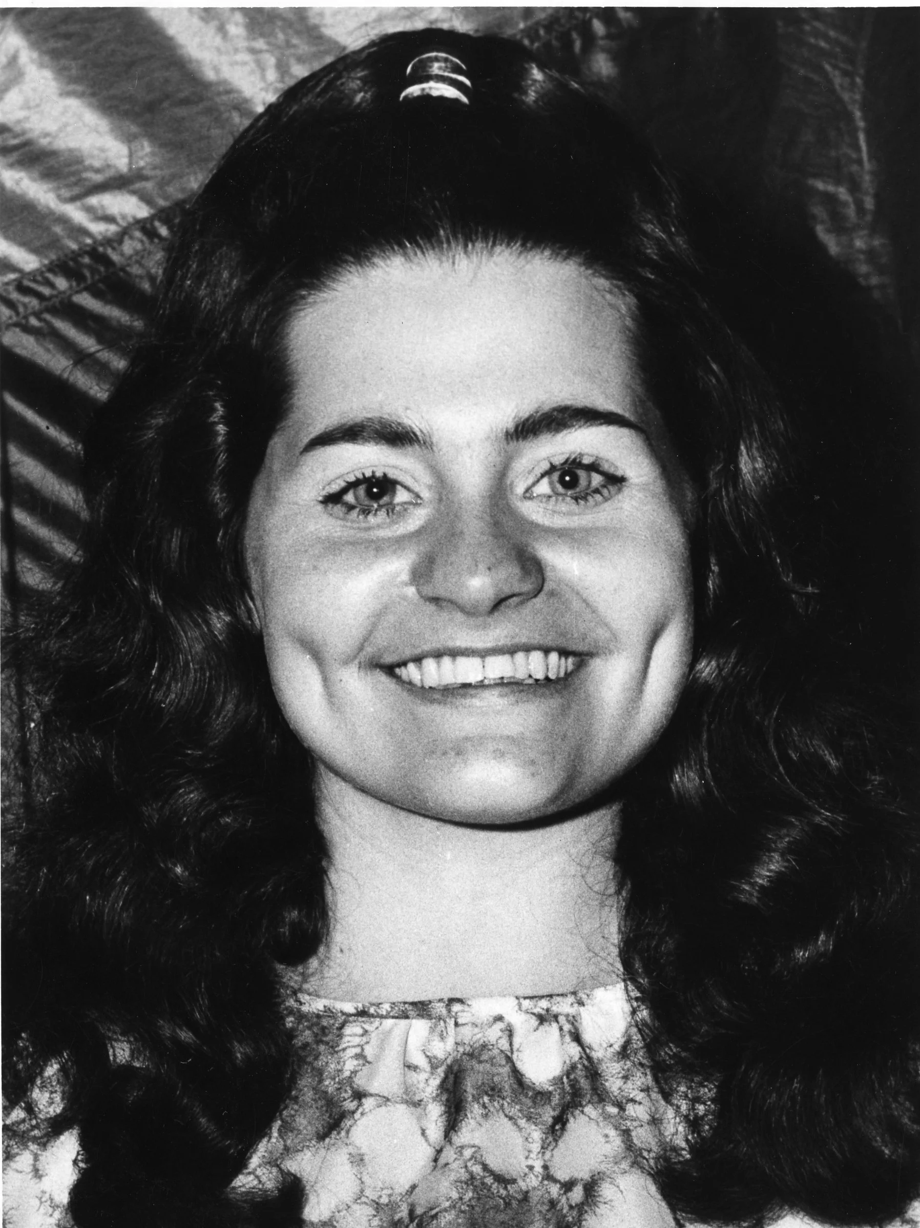 A person with long, curly hair smiling, with their face up close to the camera, and a patterned fabric or clothing visible at the bottom of the image.