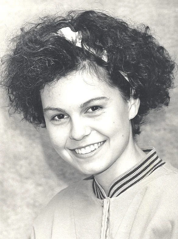 A young woman with curly hair and a headband, smiling, wearing a jacket with striped collar.