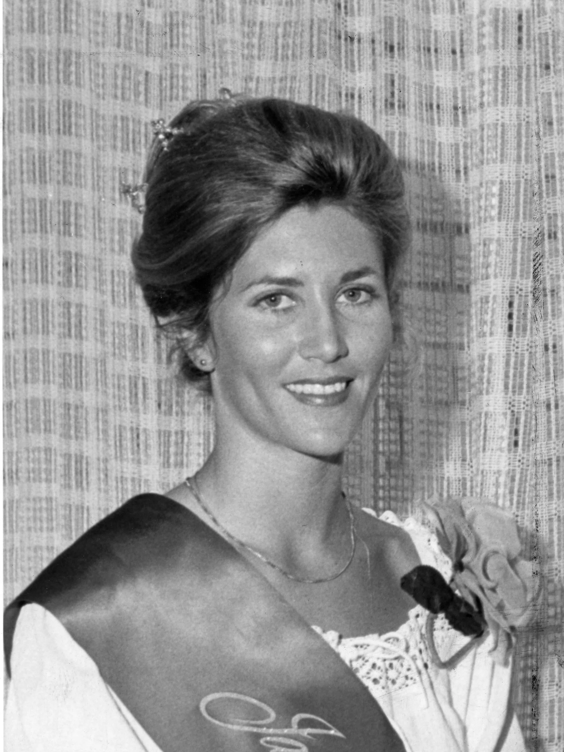 Black and white portrait of a woman with styled hair, wearing earrings, a necklace, a patterned dress, and an elegant smile, sitting in front of a textured curtain.