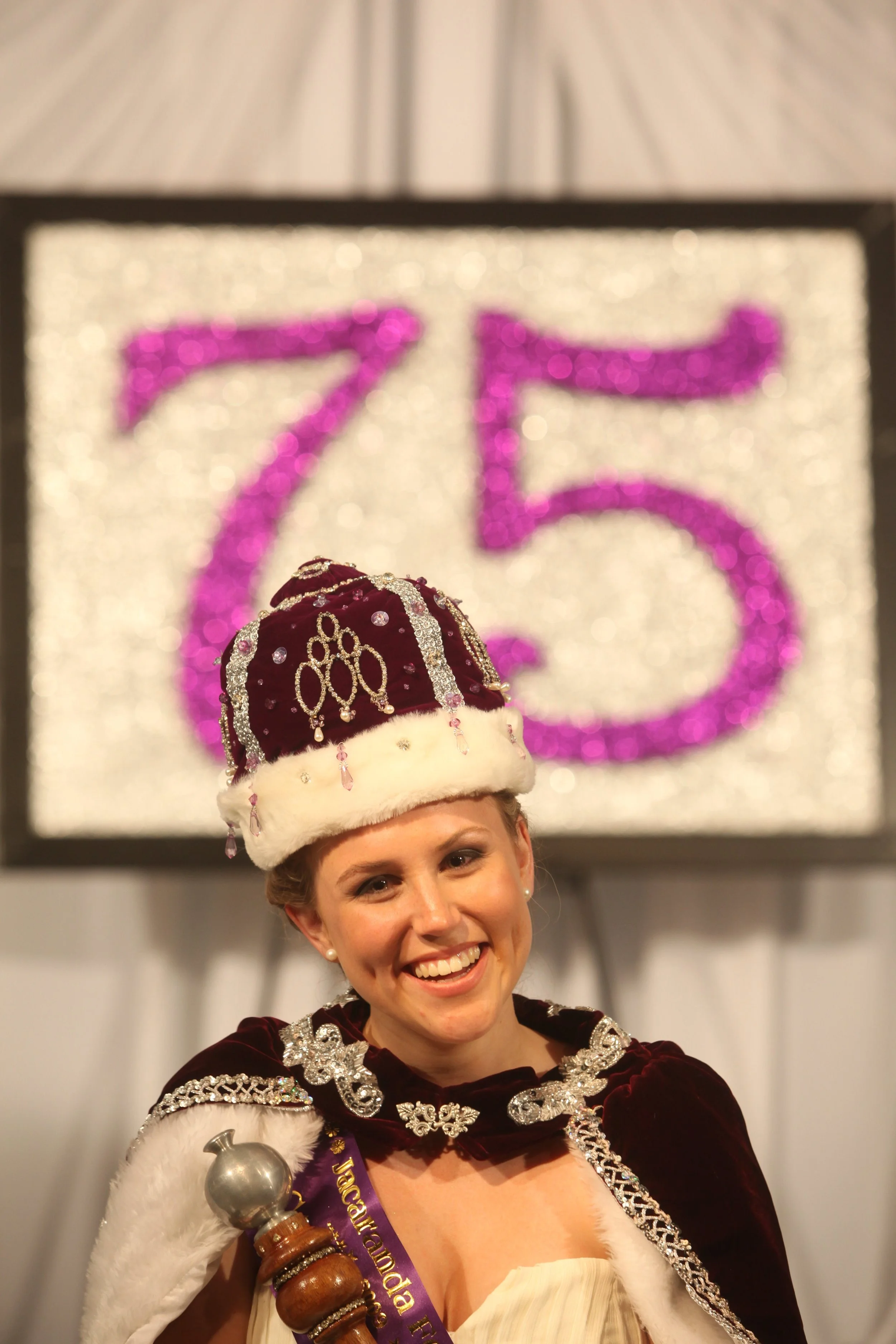 A woman dressed in a historical costume, including a crown and sash, standing in front of a glittery sign with the number 75 on it.