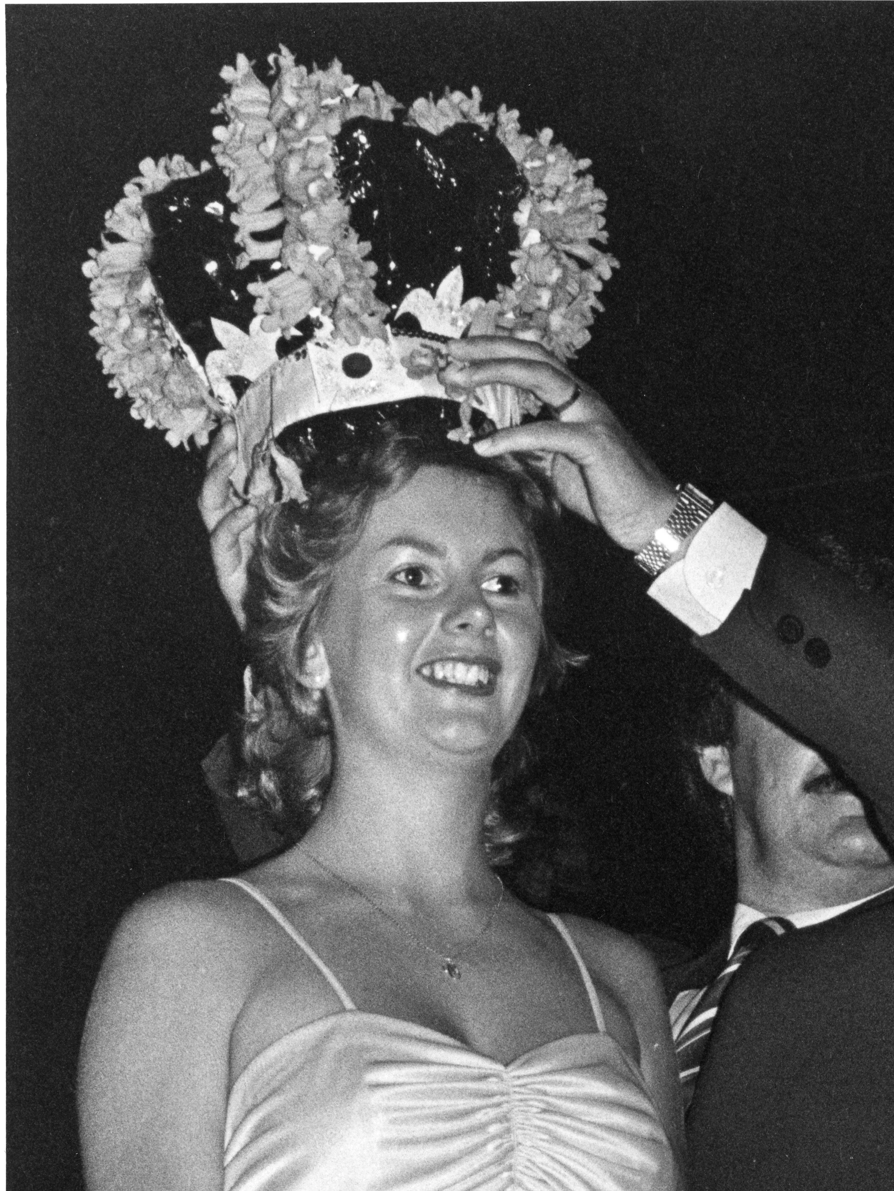 A woman wearing a dress with thin straps, smiling, with a floral headpiece made of flowers, and a tattoo on her chest.