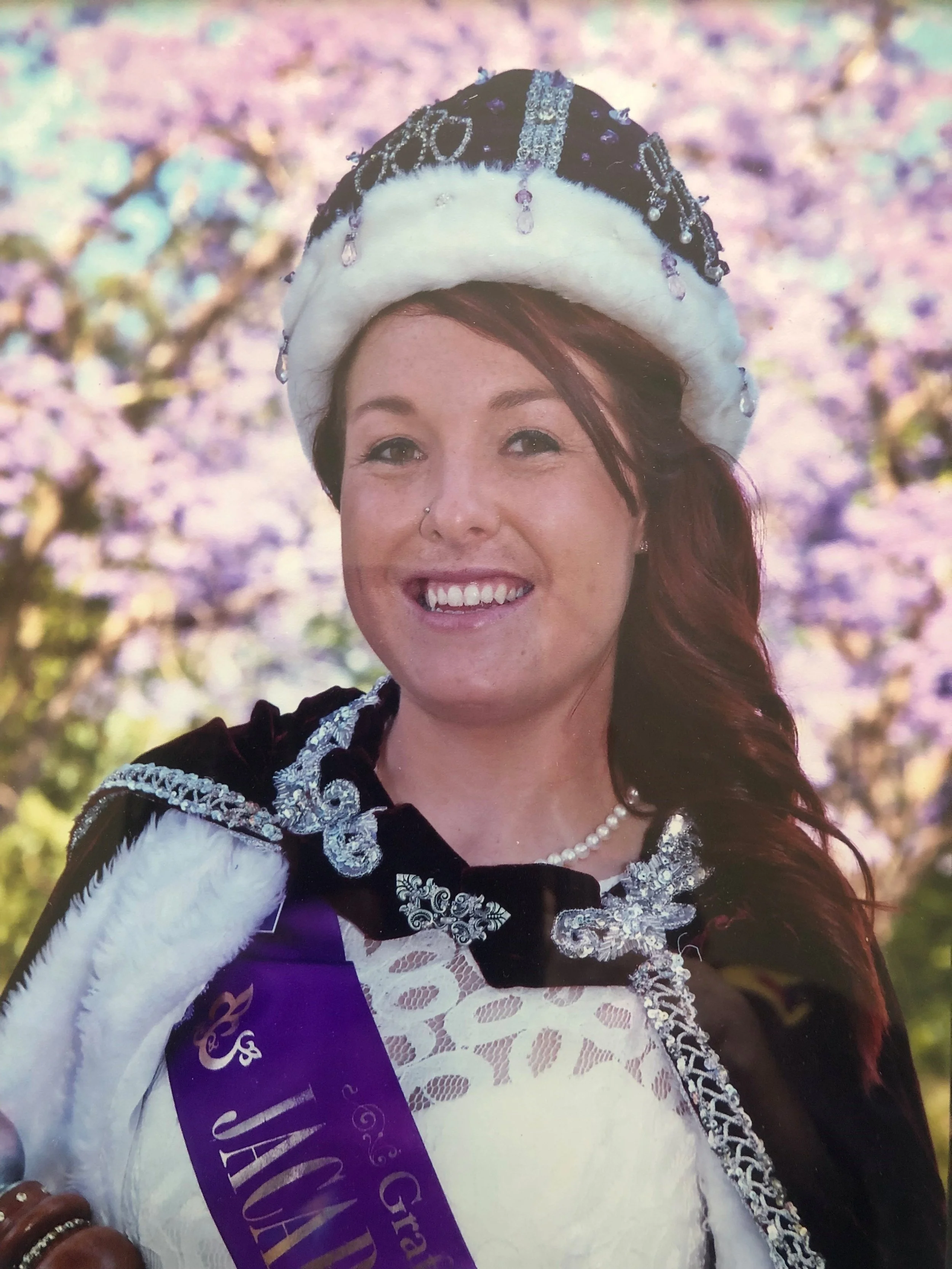 A smiling woman wearing a tiara and a sash, dressed in a fancy outfit, with a blurred background of pink cherry blossoms.
