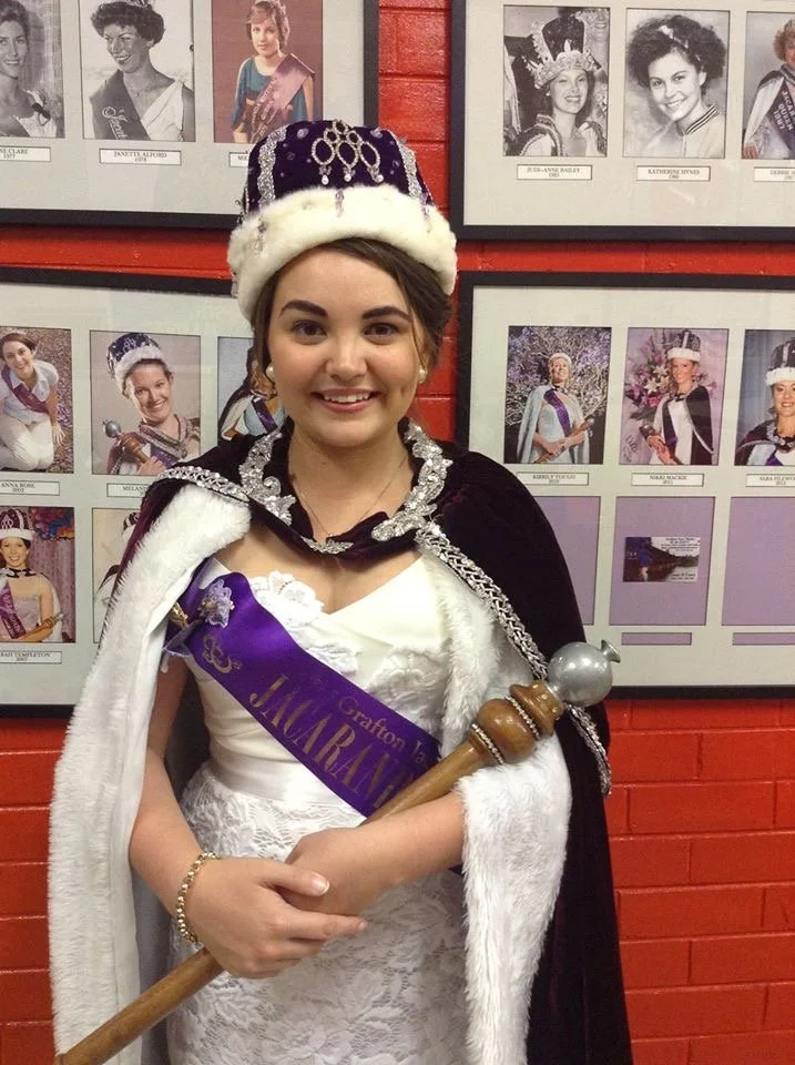 Young woman dressed in a royal costume with a crown, cape, and sash, standing against a wall with framed photos of other young women in similar attire.