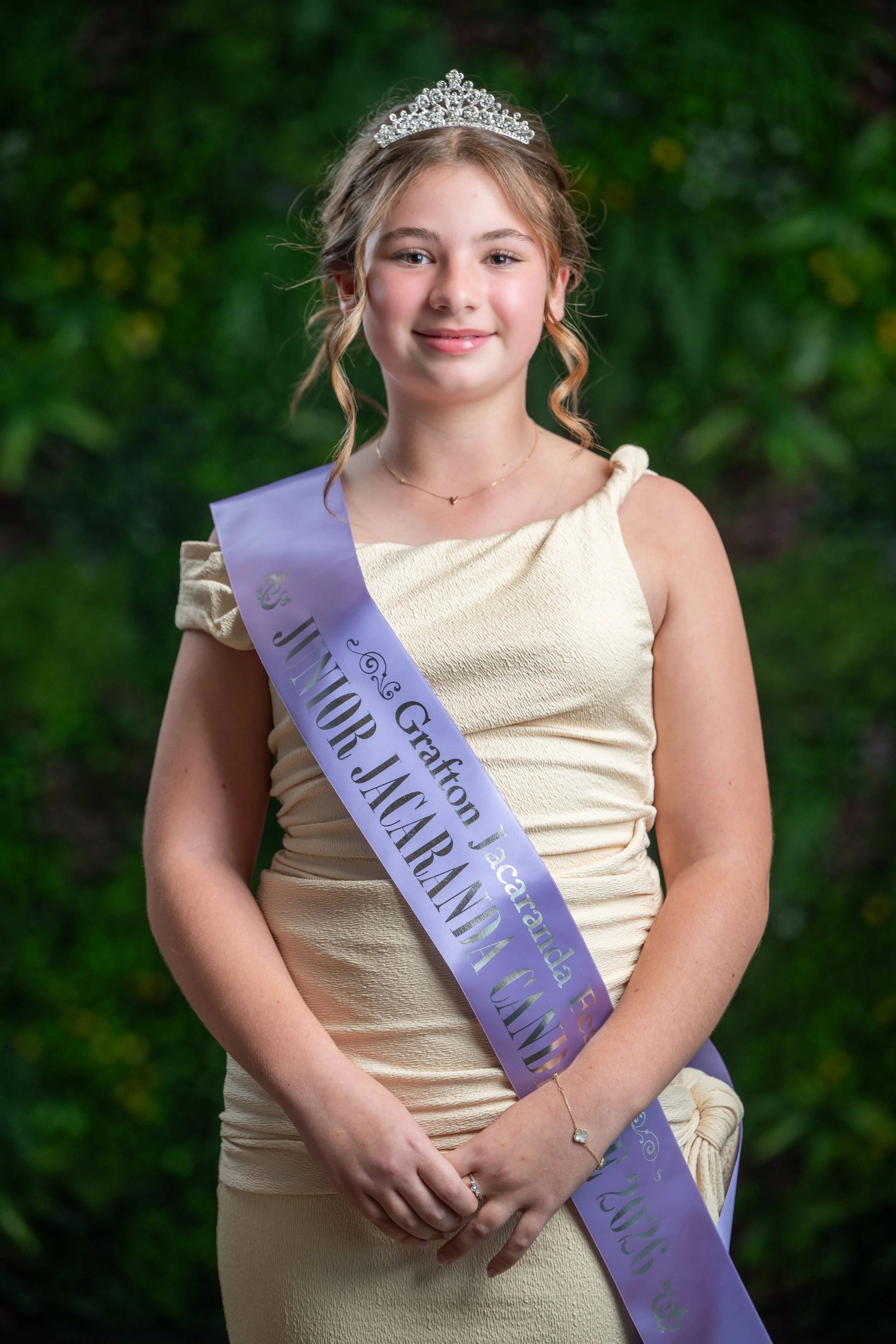 Young girl wearing a tiara and a cream-colored dress, with a purple sash bearing text, standing outdoors with green foliage background.