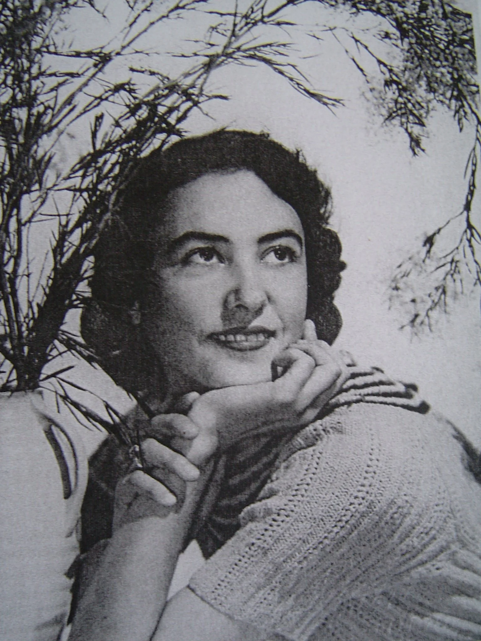 A black-and-white photograph of an Asian woman smiling, looking up and past the camera, with her hands folded near her chin. She has short, wavy hair and is wearing a textured sweater, with some tree branches visible in the background.