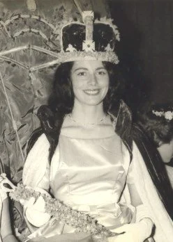 A young woman wearing a crown and a satin dress, holding a bouquet, at a celebration or pageant event.