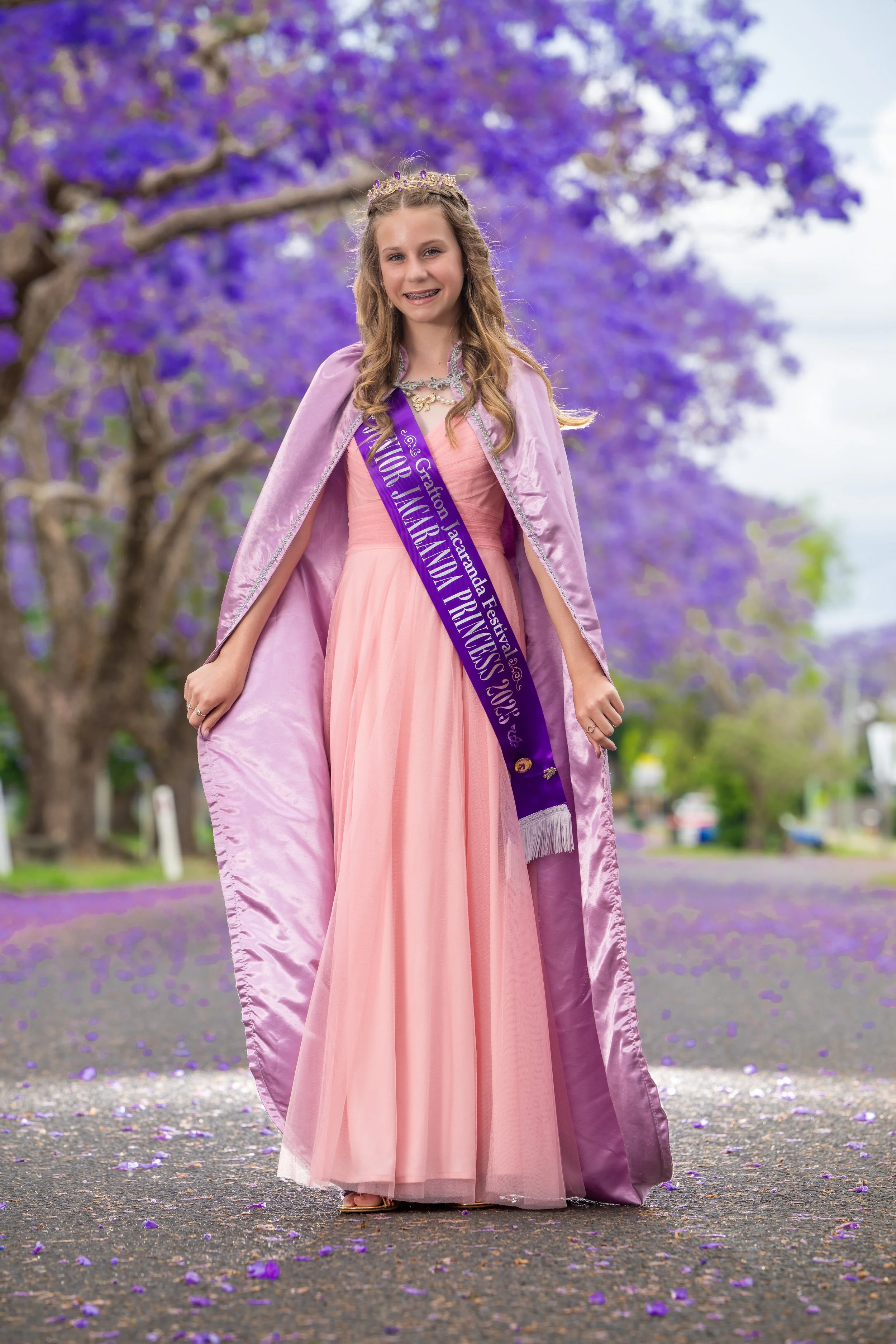 Young girl dressed as a princess wearing a pink dress, purple sash, and tiara, standing on a street with purple flowers from a blooming tree in the background and purple petals on the ground.