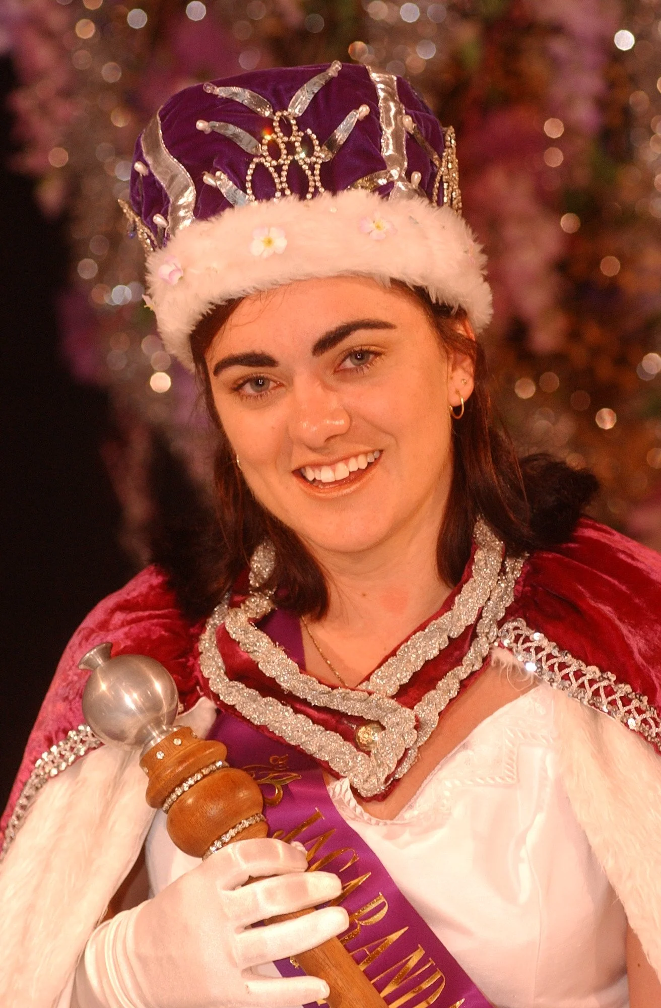 A woman smiling, wearing a purple and white crown with gold embellishments, dressed in a festive costume with white gloves, red velvet cape with silver trim, and holding a scepter, with a background of pink and purple lights.