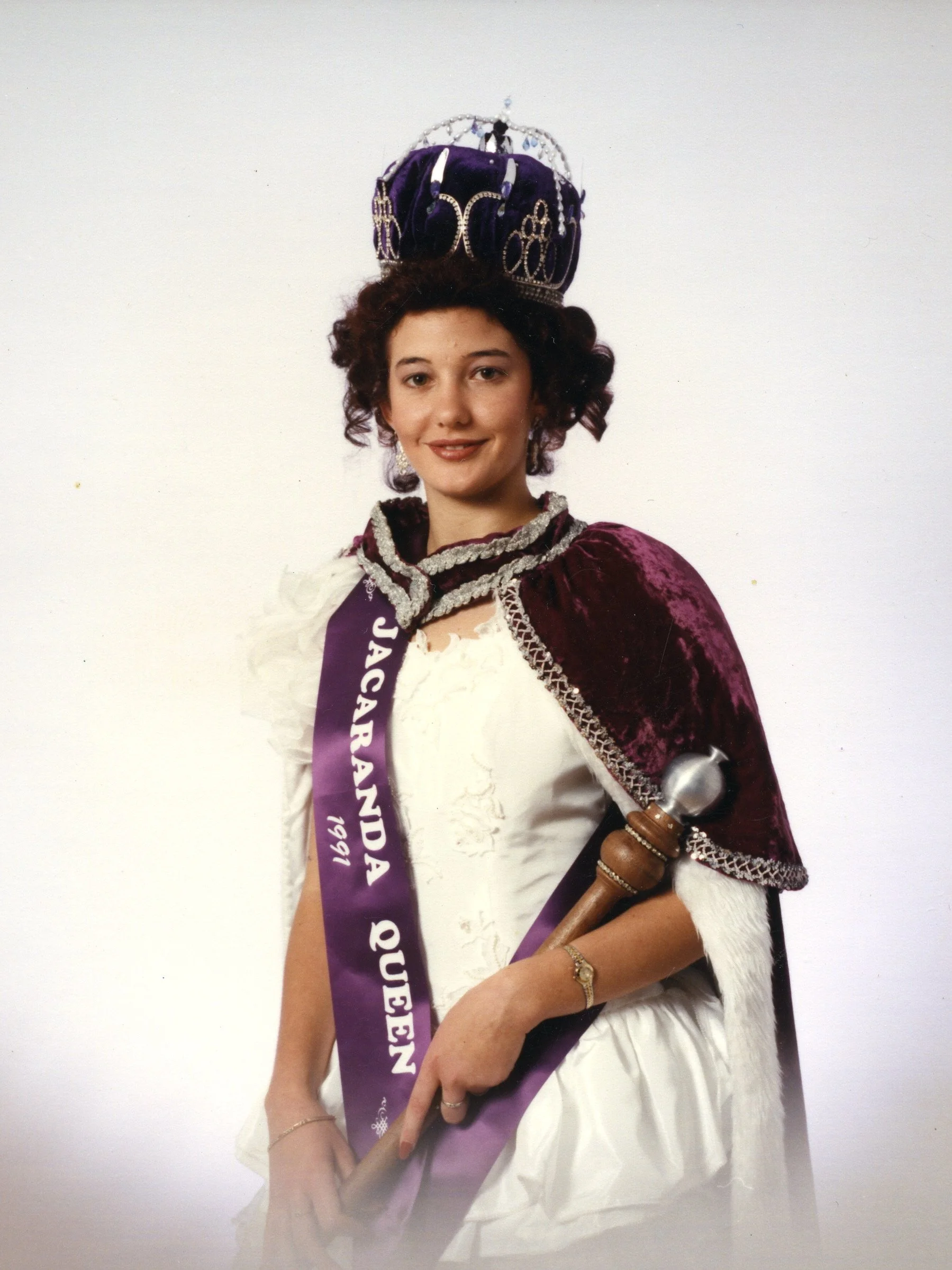 A woman dressed in a white gown with a maroon velvet cape, wearing a crown and a sash, holding a wooden scepter with a silver top, posing for a photo against a plain white background.