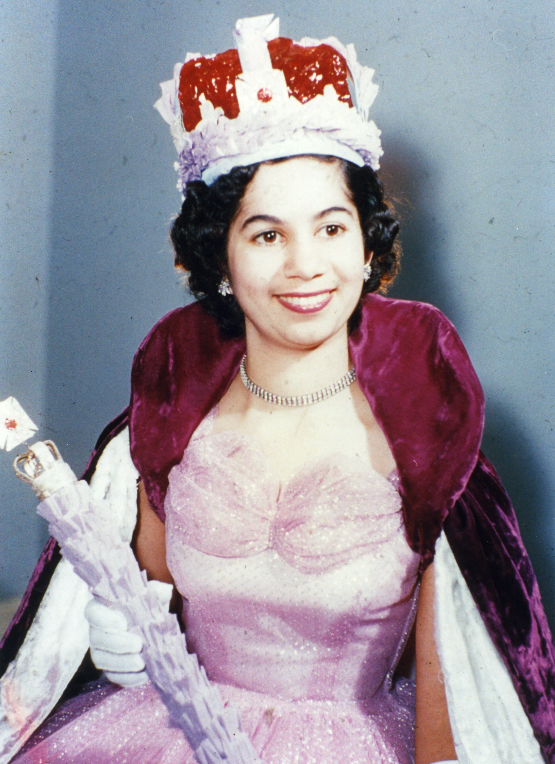 A woman dressed as a princess, wearing a pink gown, white gloves, a crown, and a purple cape, smiling at the camera.
