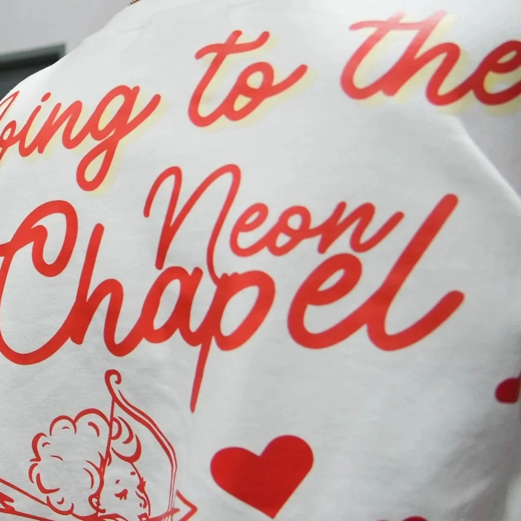 Close-up of a shirt with red cursive text and heart illustrations, partially showing the phrase 'Neon Chapel' and "going to the Neon Chapel"