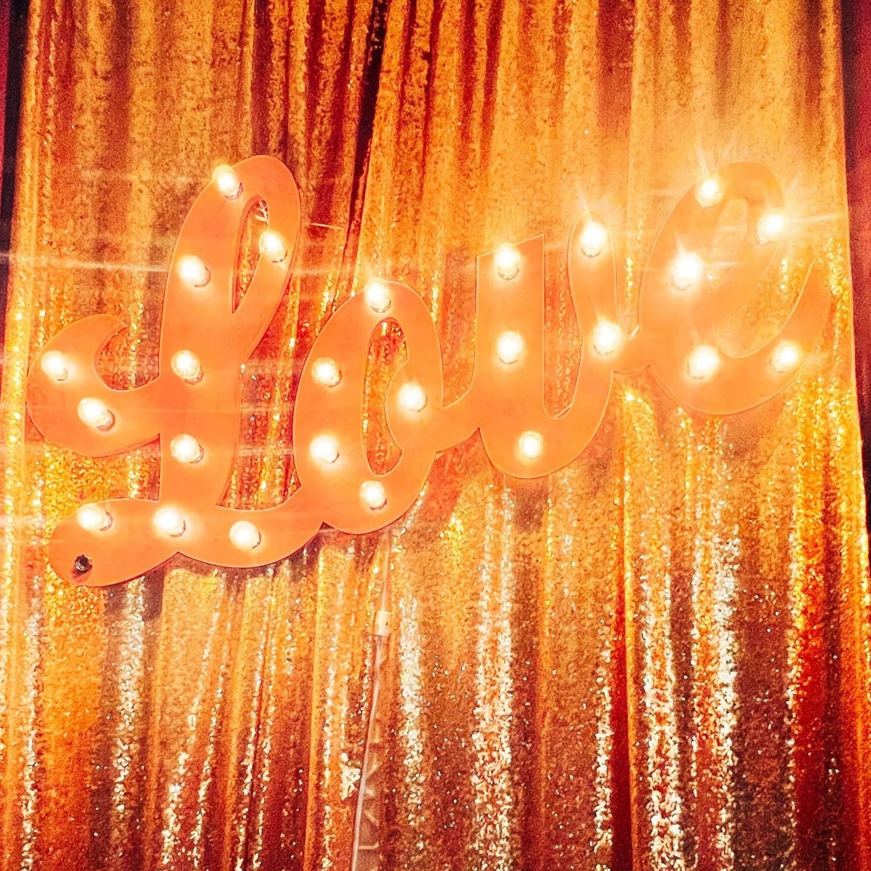 A illuminated sign spelling 'love' in cursive script, with small light bulbs, mounted on a golden, glittery curtain background.