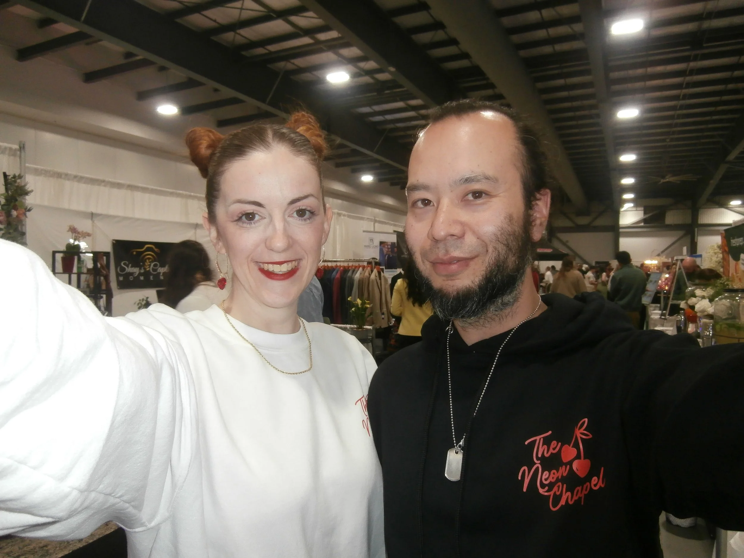 A man and woman taking a selfie at an indoor event or market, with clothing racks and other people in the background.