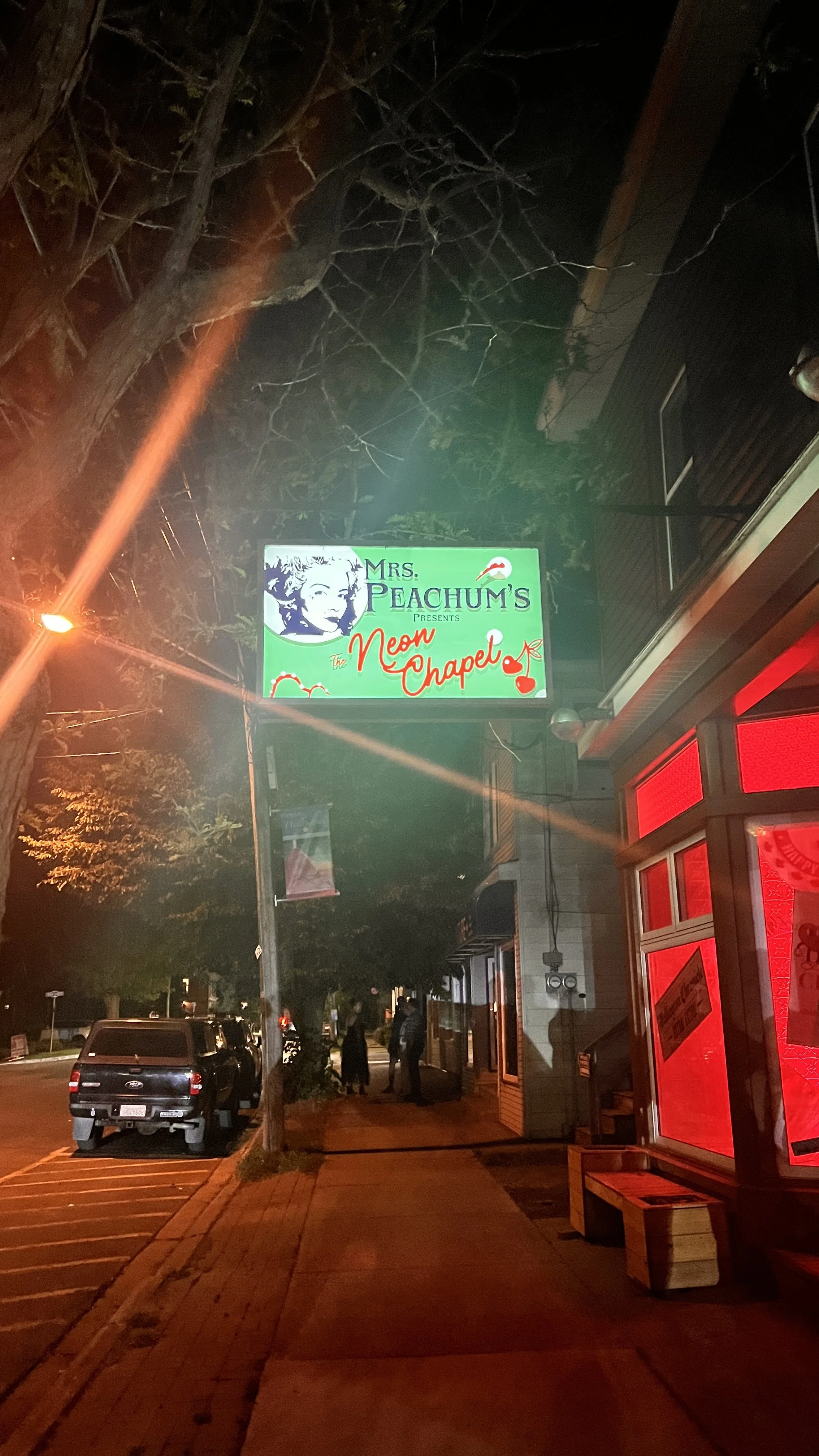 Night time street scene in Prince Edward County with a lit sign reading 'Mrs. Peachum's presents The Neon Chapel,' red neon-lit storefront, parked black pickup truck, pedestrians, and trees overhead.  