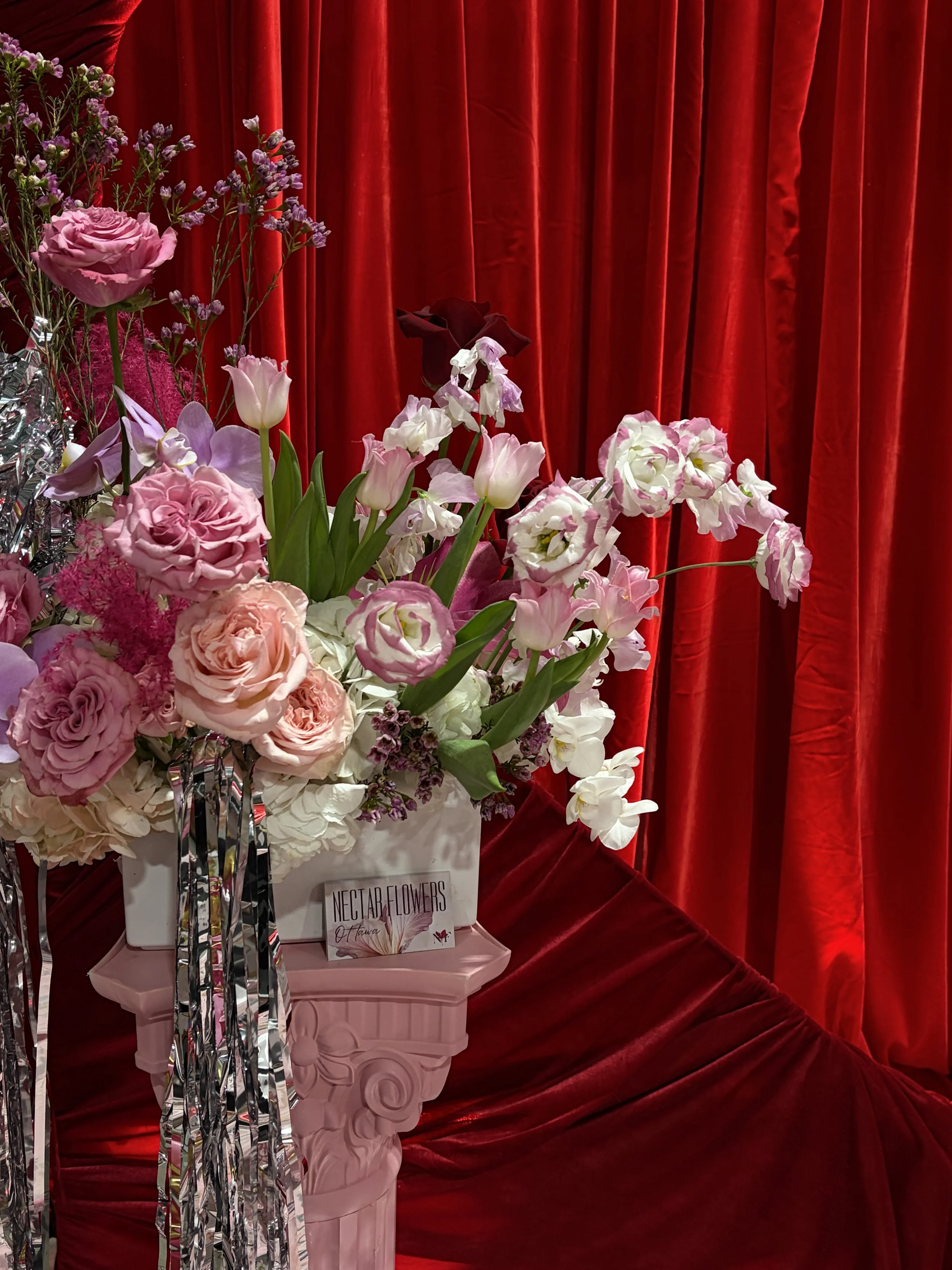 A bouquet of pink and white flowers in a pink vase on a pink table, with a red velvet curtain in the background.