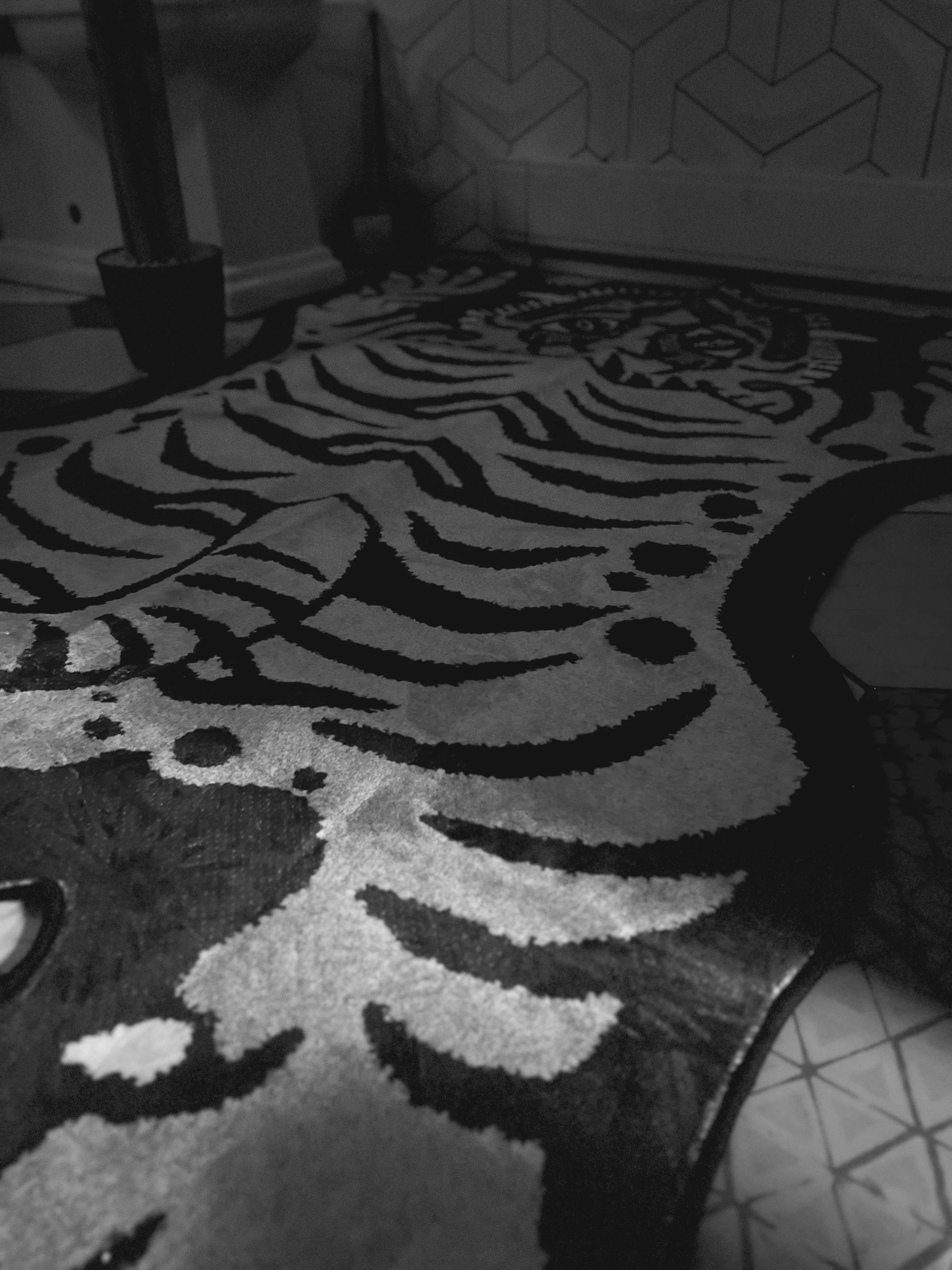 A black and white patterned rug on the floor with a tiger face design, partially visible. There is a potted plant with a curved stem to the left and a wall with a geometric pattern in the background. Part of a tiled floor is visible on the right side