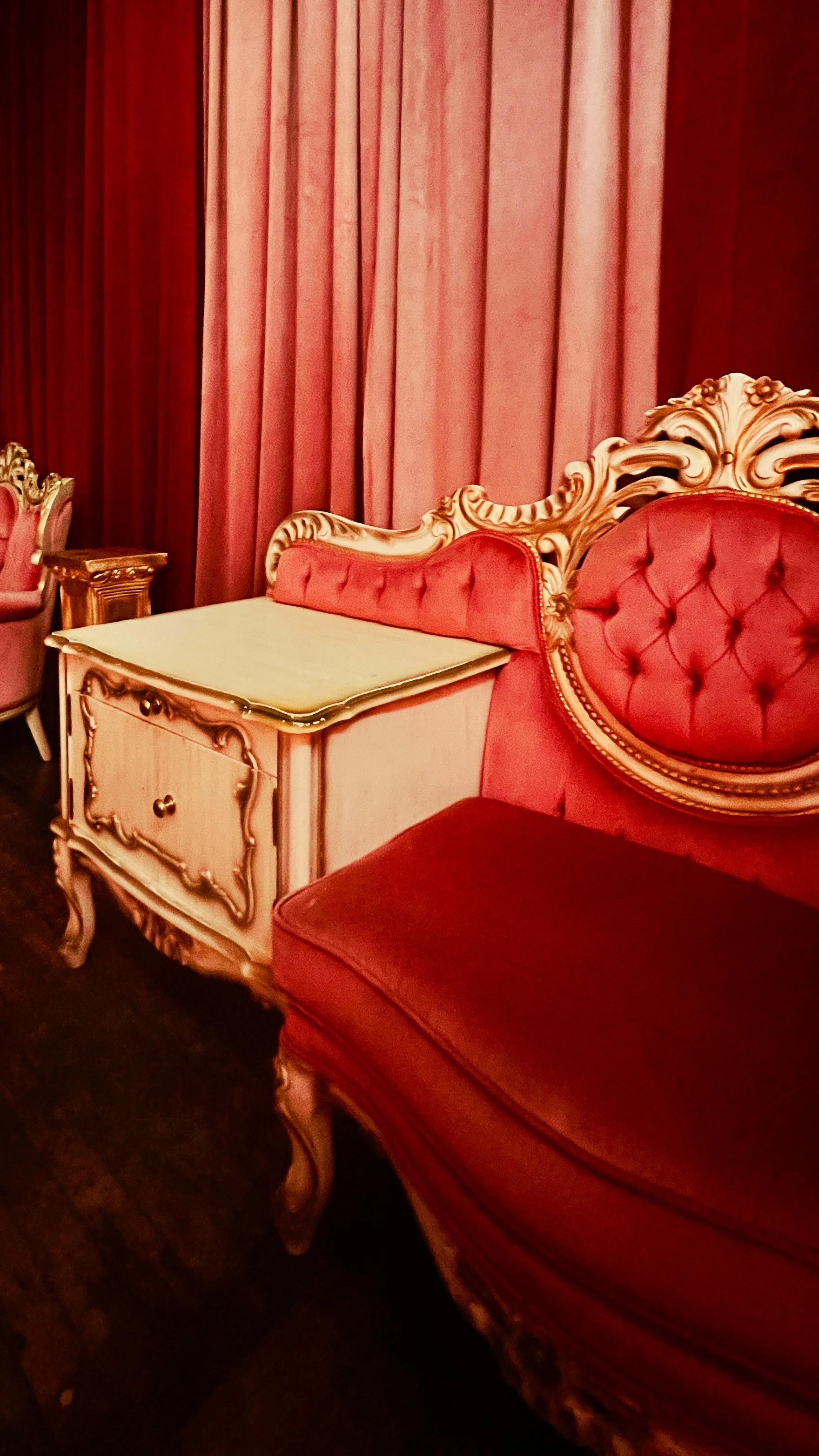 A vintage pink and gold ornate couch with a tufted backrest, next to a small white and gold side table, set against pink curtains in a luxurious room.