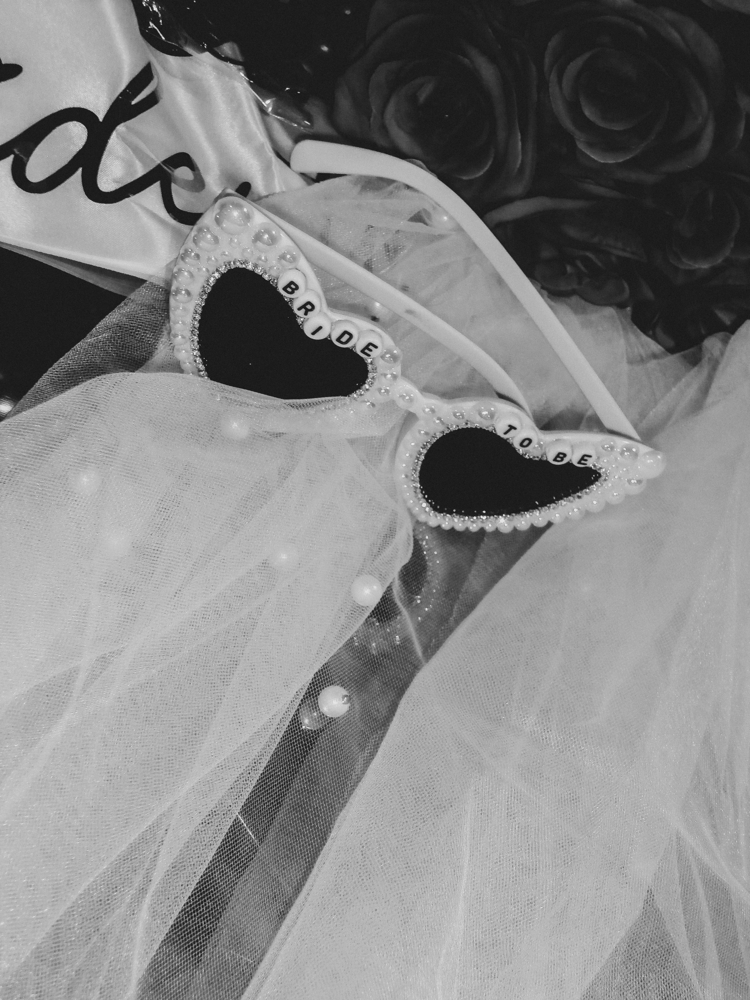 Close-up of decorative items including two heart-shaped accessories with pearls and words, a bouquet of roses, and a gift with a ribbon, in black and white for a Bride.