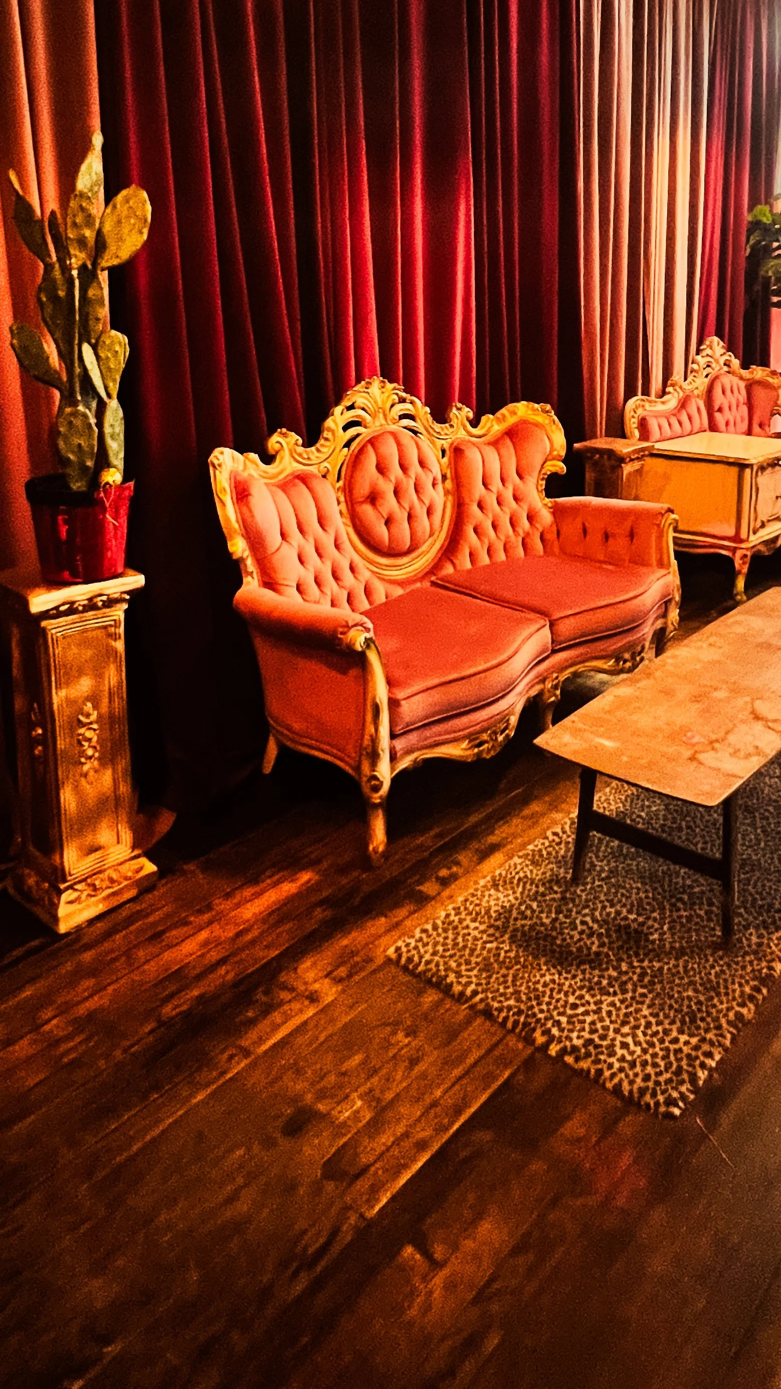 Vintage pink velvet loveseat with ornate gold trim, set against deep red curtains, with a potted plant on a decorated pedestal to the left and a wooden coffee table with a leopard print rug underneath.
