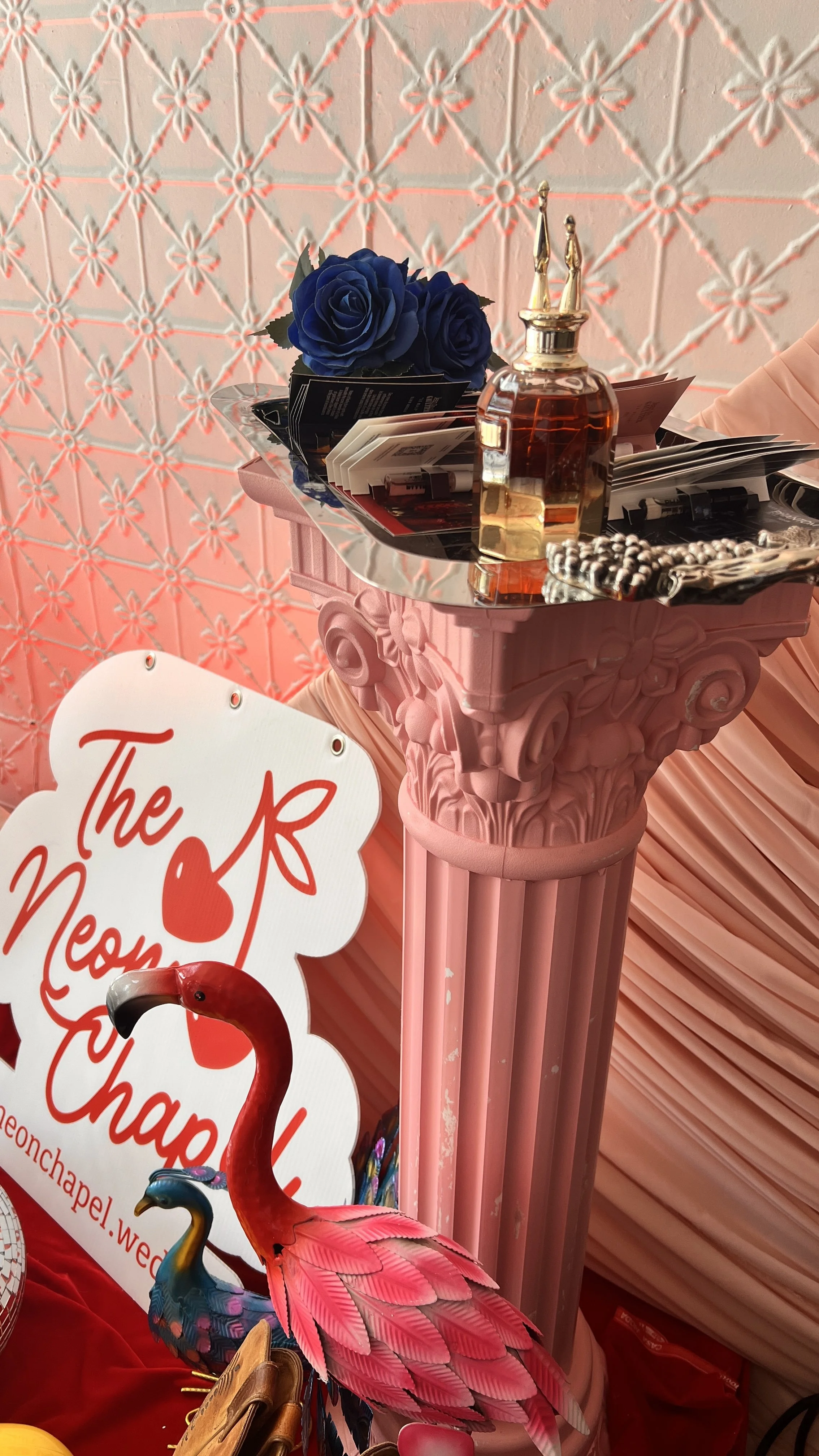 Decorative pink classical column with ornate capital, topped with a silver tray holding blue roses, brochures, and a small perfume bottle. In front are pink flamingo and peacock figurines, and a sign with red text reading 'The Neon'. The background f