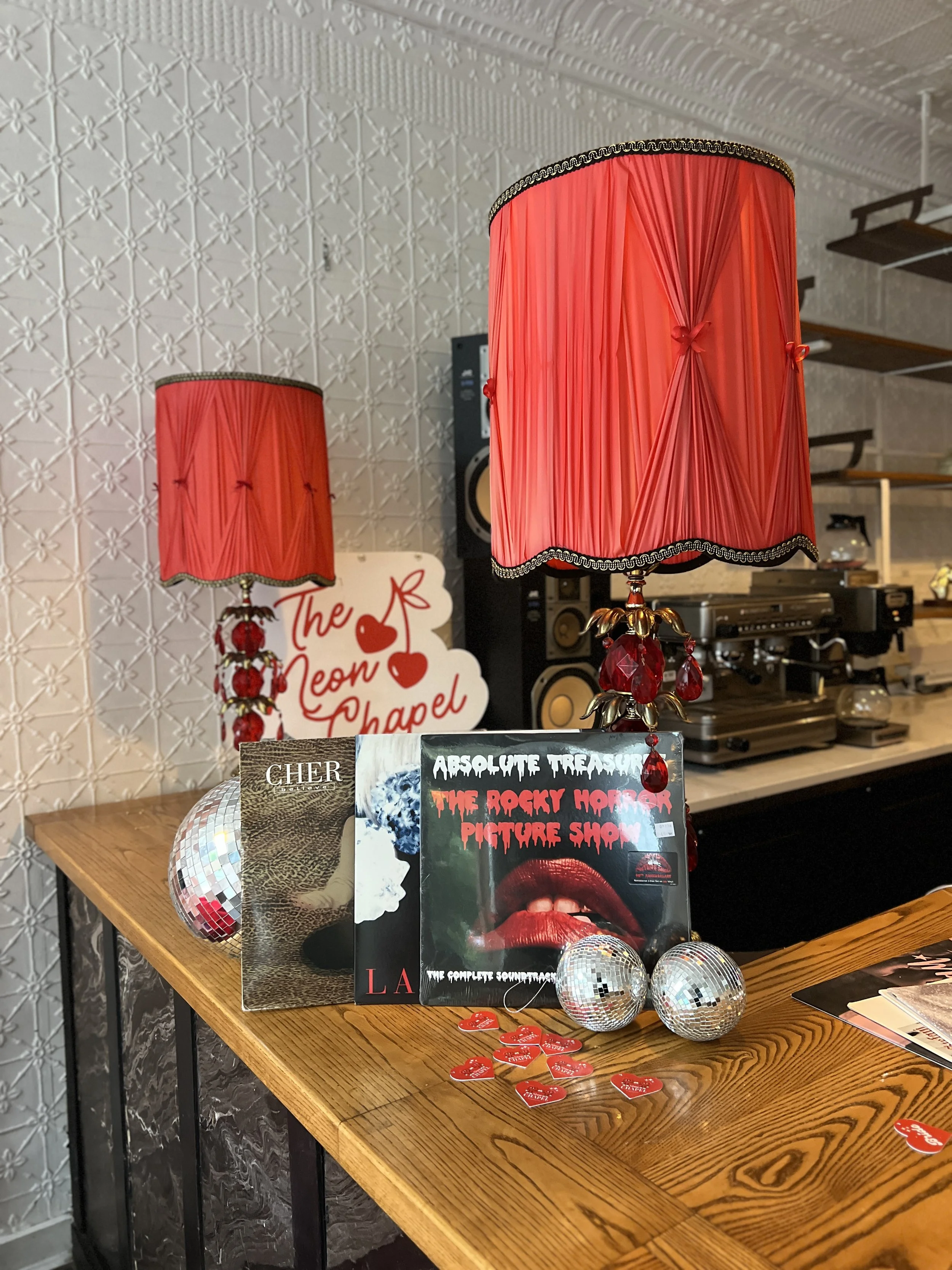 Two red table lamps with ornate bases, placed on a wooden table. Surrounding them are promotional booklets, small red heart-shaped items, and decorative disco balls. In the background, there is a textured wall with floral patterns, a sign that reads 