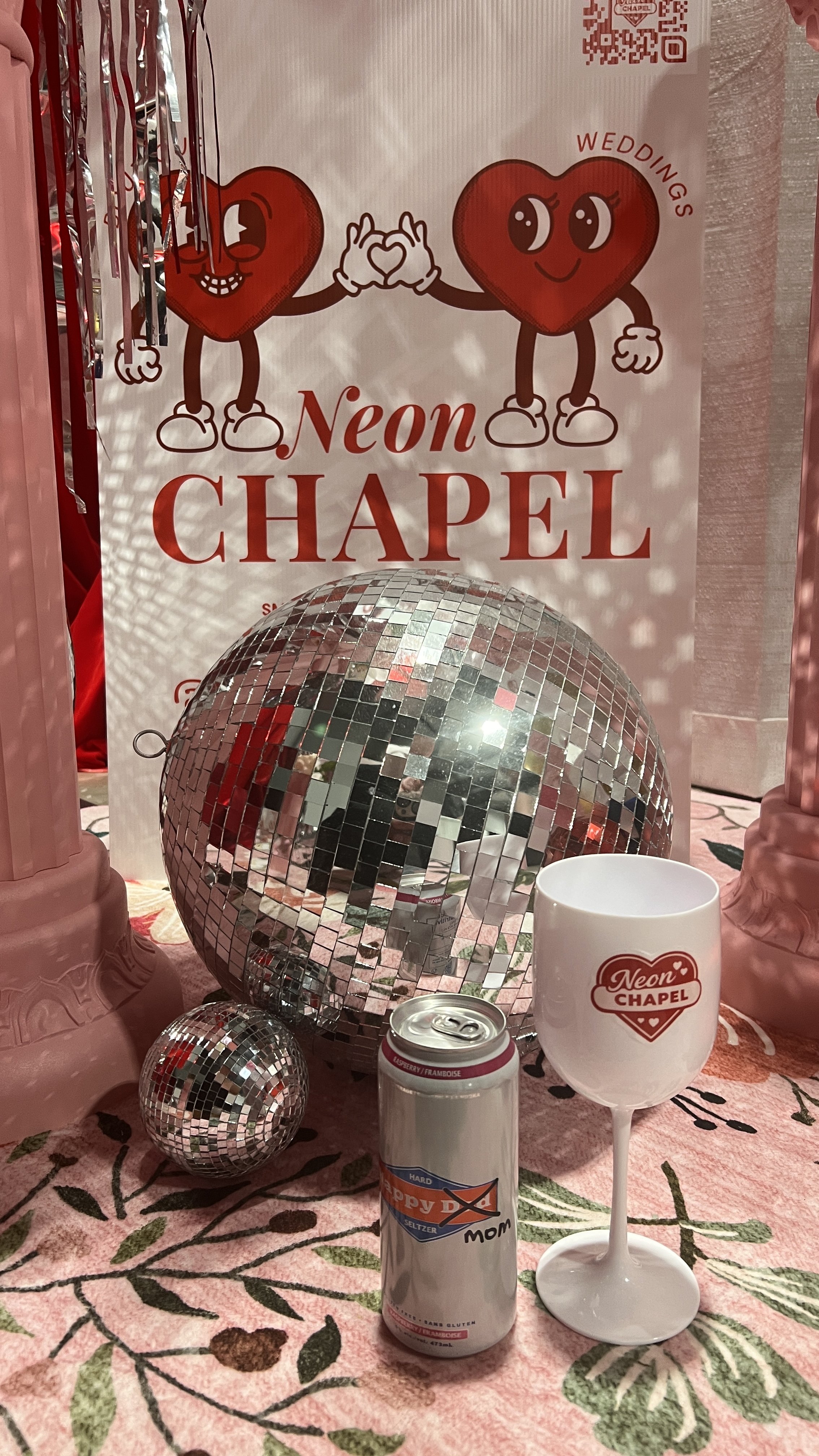 Decorative table setup for a wedding or celebration with a sign reading 'Neon Chapel' featuring cartoon hearts in the background. There is a shiny disco ball, a white cup with a 'Neon Chapel' logo, a soda can, and a Seltzer water bottle on a pink and