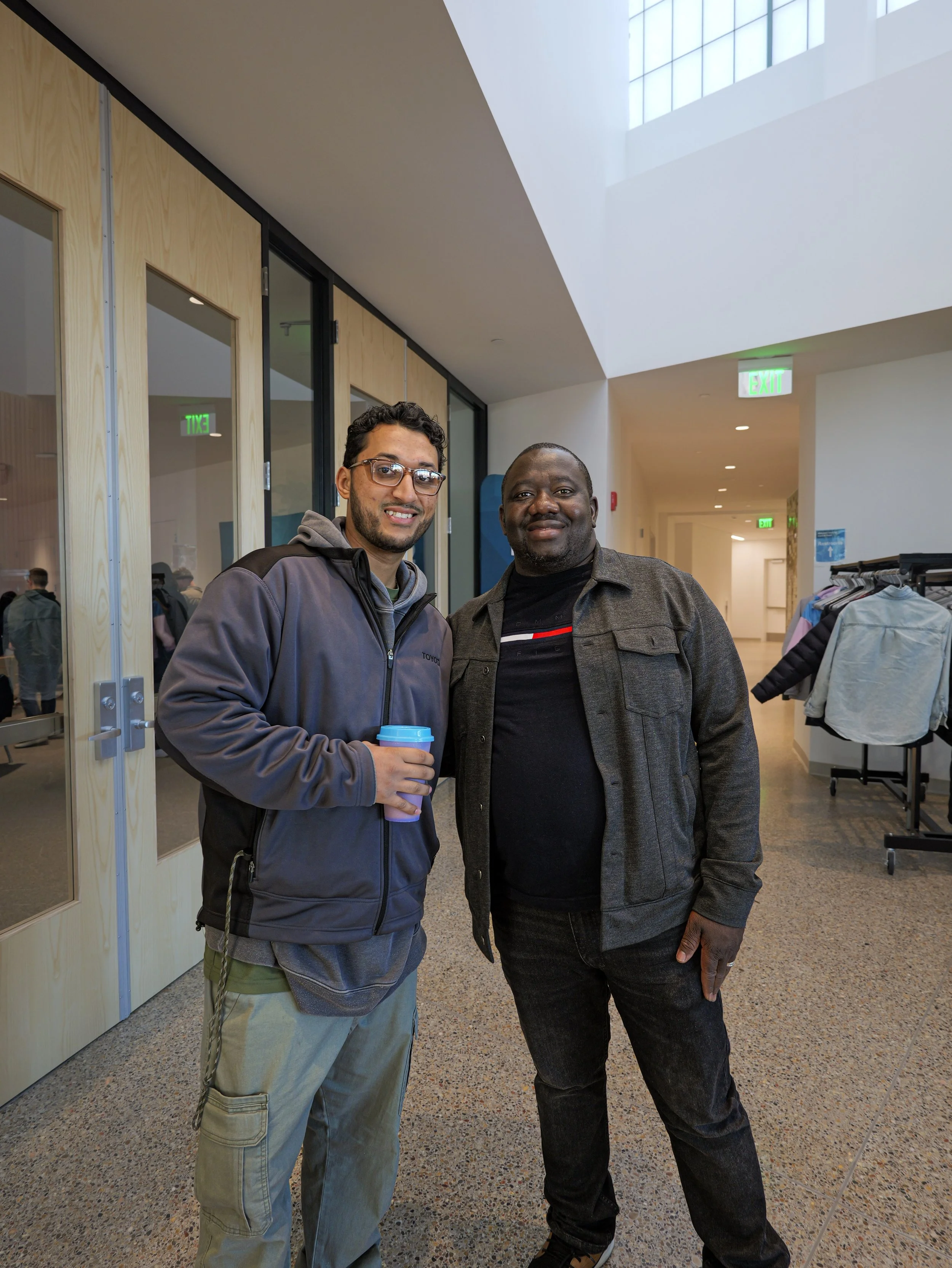 Two men standing indoors, smiling, with one holding a coffee cup. They are in a modern church in the Twin Cities with glass doors, wooden wall accents, and happy people.
