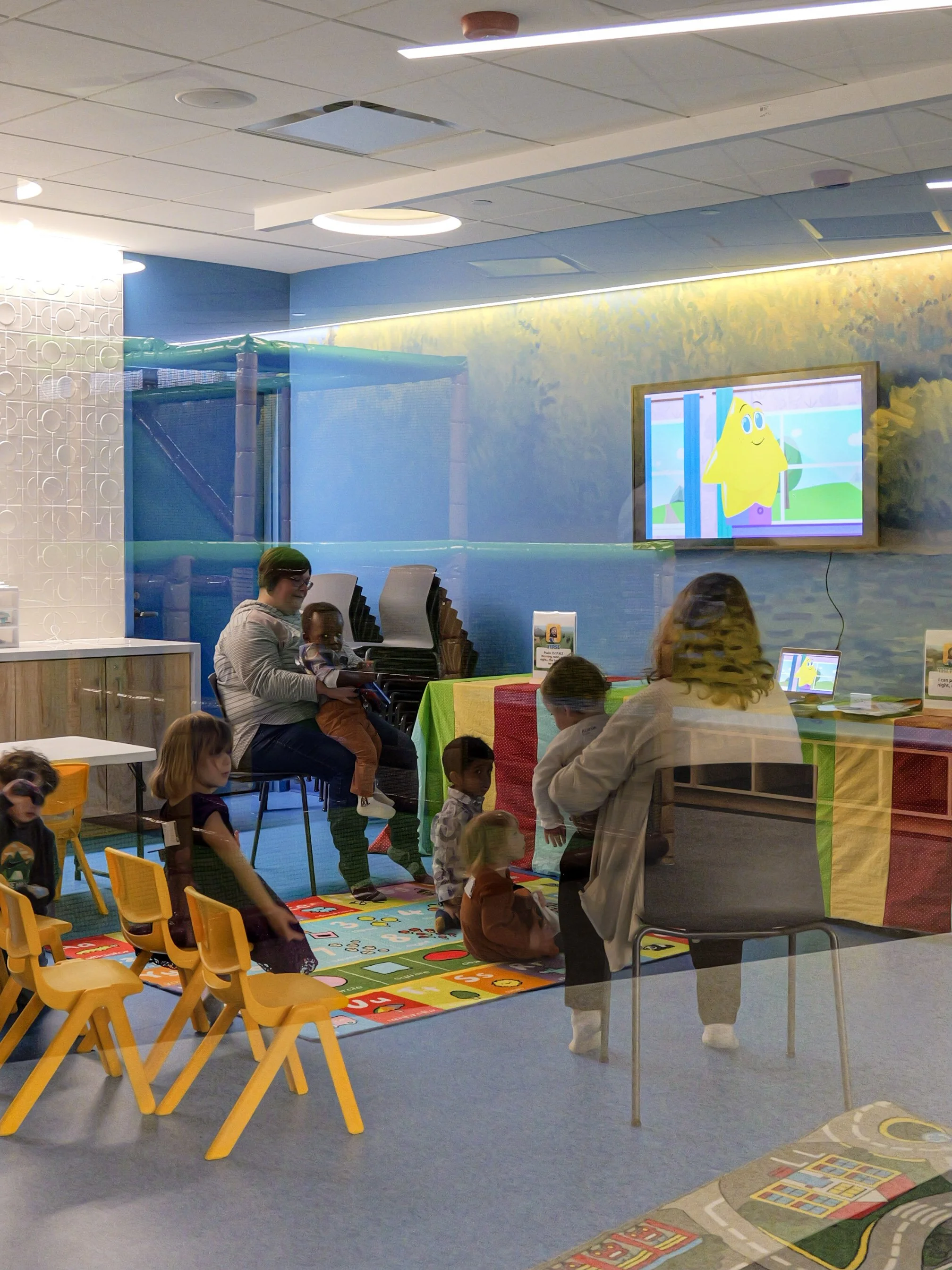Children and adults in a play area at a church for families with colorful rugs, toys, and tables, laughing  and engaging in activities.