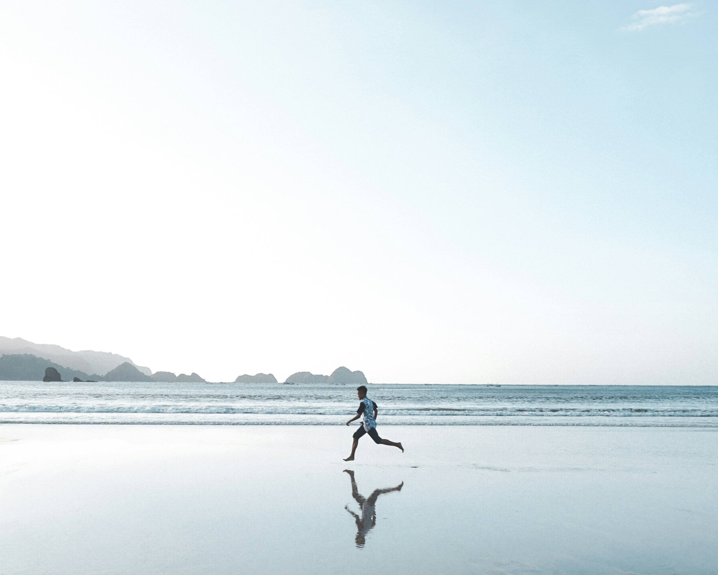 A person running on a beach near the ocean with mountains in the background and a large body of water reflecting the scene.