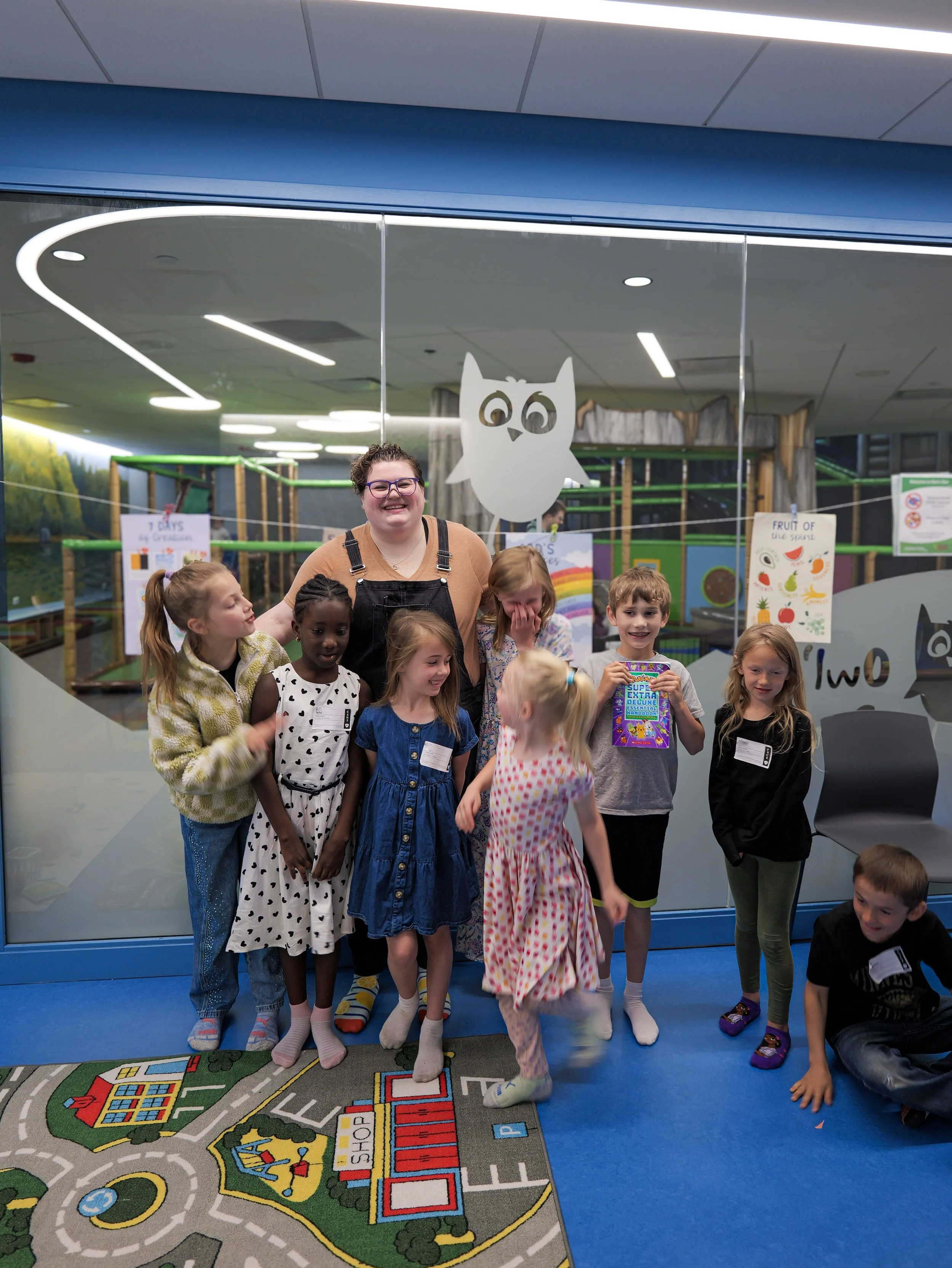 Children and an adult standing on a colorful children's play carpet, inside a building with a glass wall with an owl graphic, indicating an indoor play or educational area.