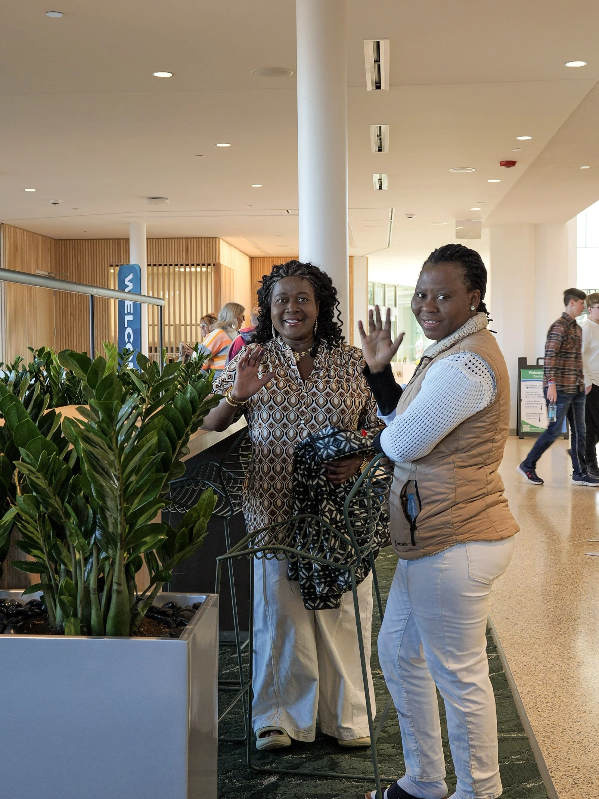 Two women are standing and smiling at Sunday church service Woodbury, waving at the camera. There is a table with a green plant in front of them. Several people are walking and talking in the background.